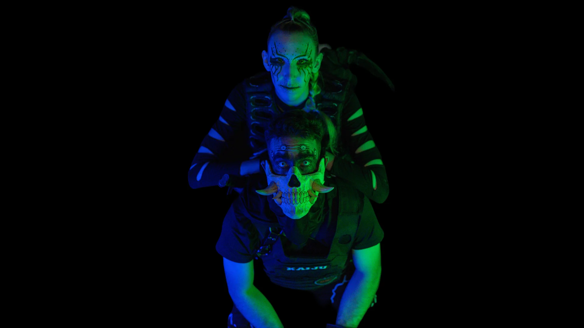 Two people in dark clothing and face paint pose against a black background, with one person standing behind the other and placing their hands on the other’s head. Green and blue lighting highlights their faces.