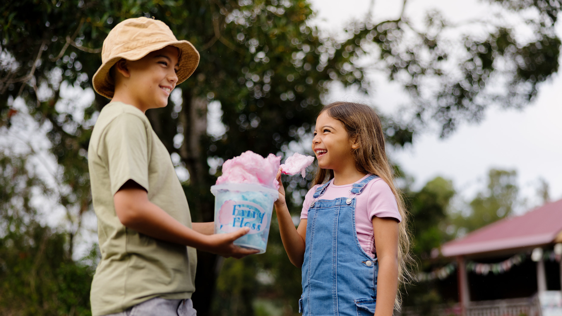 Two children smile at each other outdoors; one holds a bucket labeled “Fairy Floss” filled with pink cotton candy, while the other, wearing overalls, holds a piece of the candy. Trees and a house are in the background.