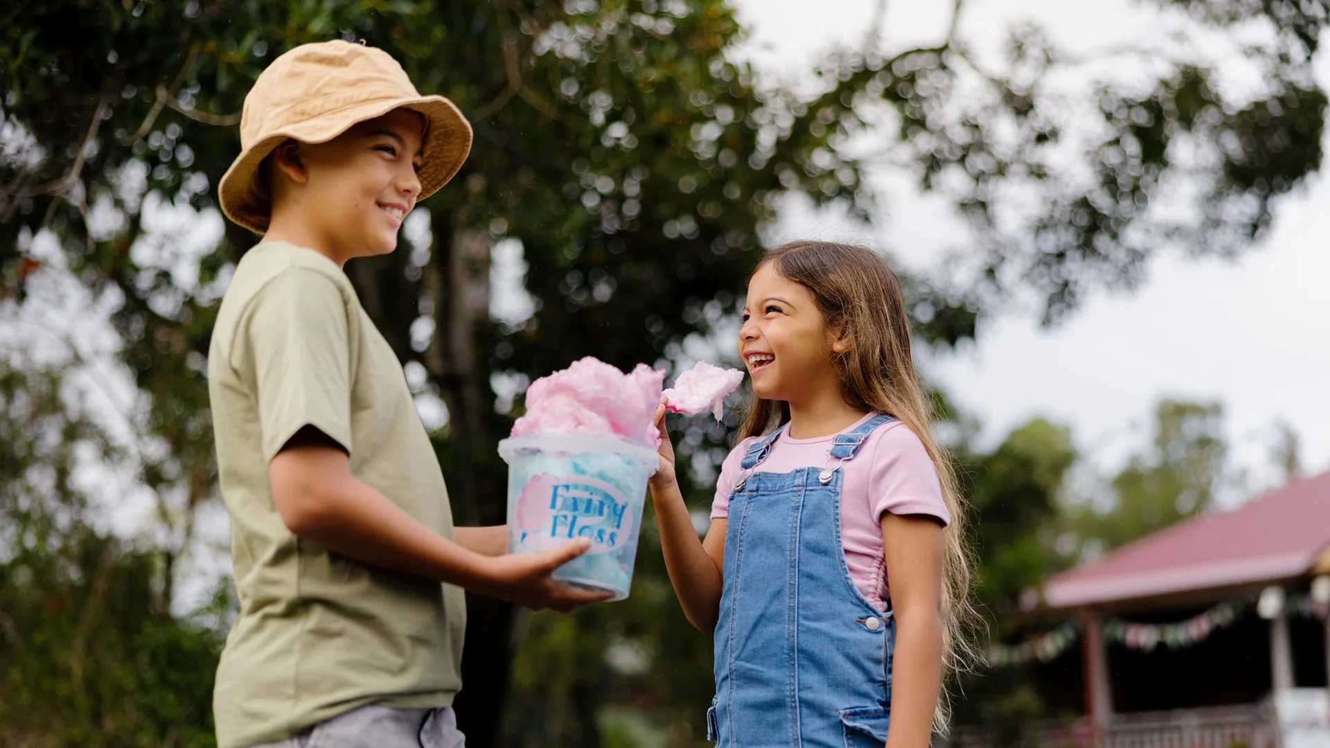 Two children smile at each other outdoors; one holds a bucket labeled “Fairy Floss” filled with pink cotton candy, while the other, wearing overalls, holds a piece of the candy. Trees and a house are in the background.