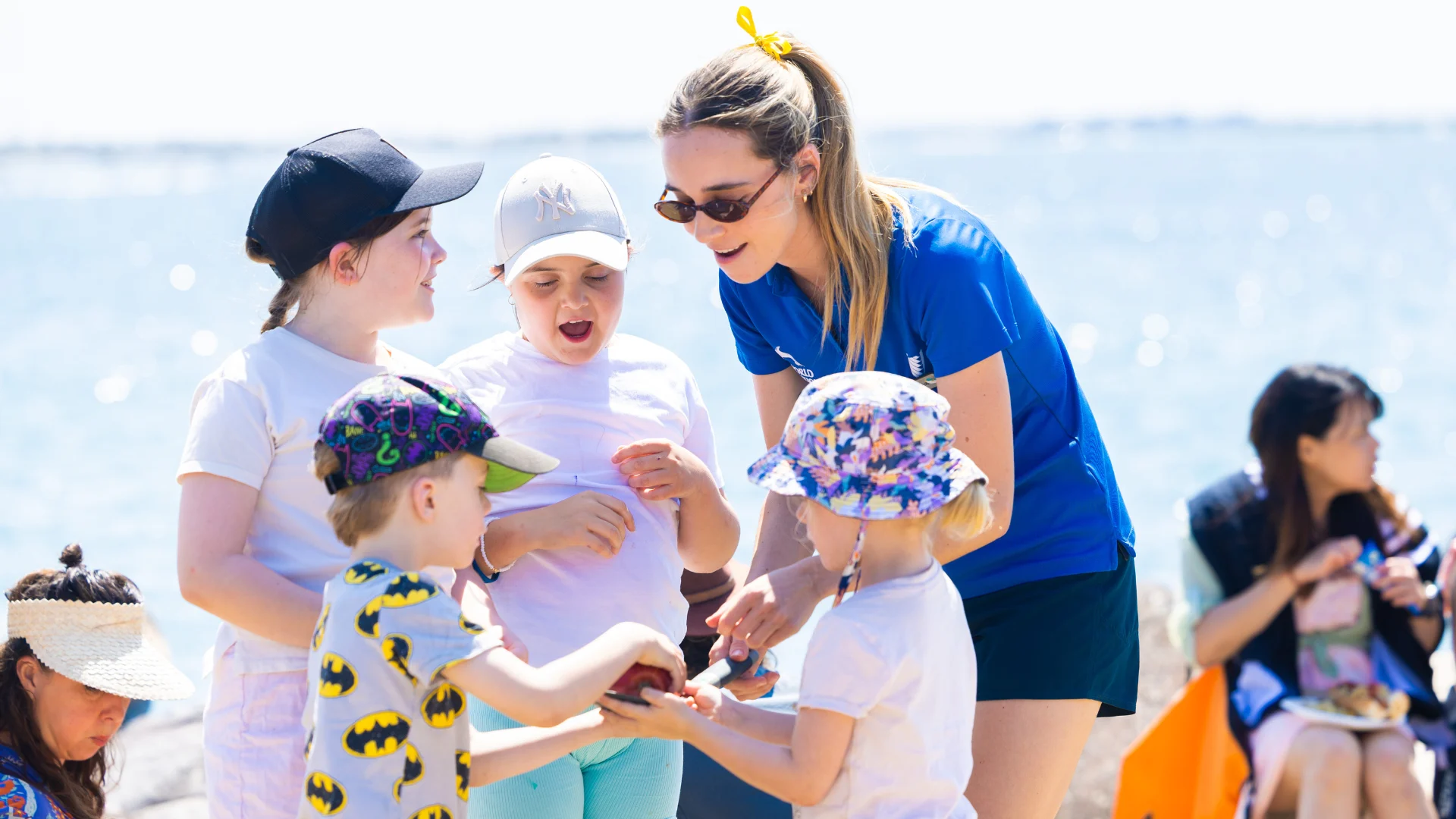 A group of children and a woman wearing sunglasses stand together outdoors by the water on a sunny day, smiling and interacting. Other people are sitting and talking in the background.