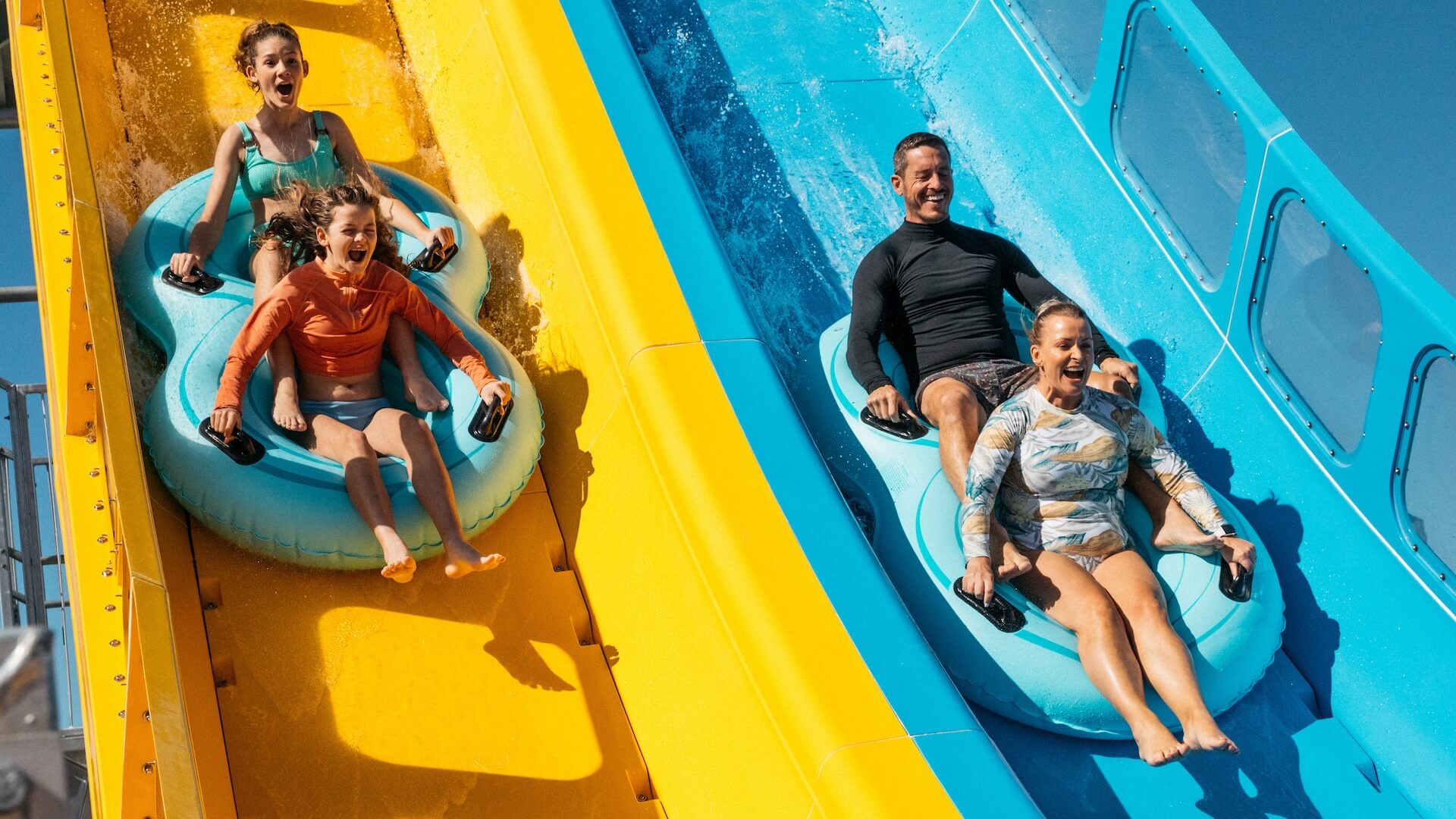 Family enjoying the Kaboom! waterslide at Wet’n’Wild Gold Coast, racing side by side on bright yellow and blue slides with inflatable rafts for a thrilling water park experience