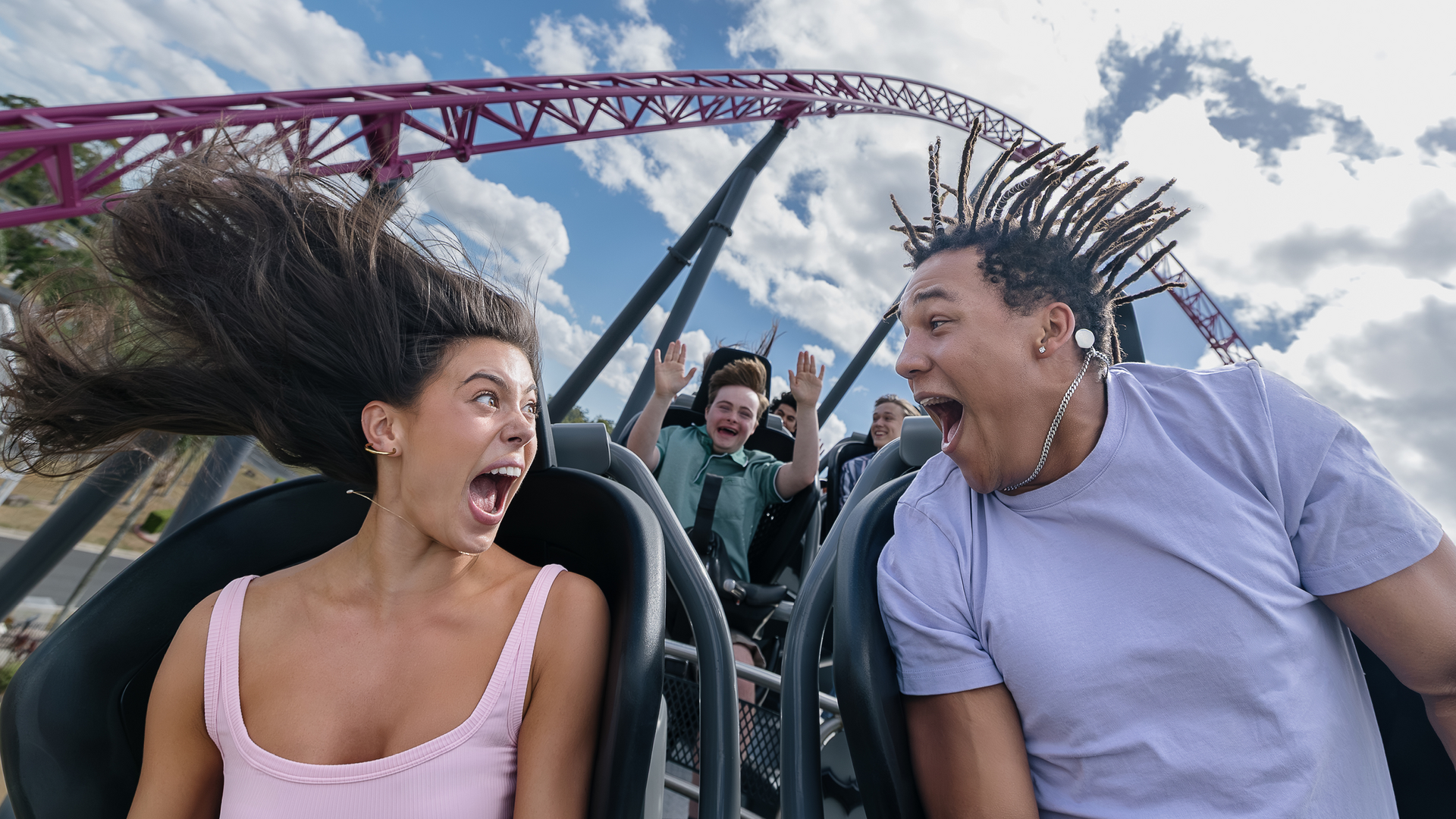 Two people in the front seats of a roller coaster scream with excitement, hair blown back, while two more riders behind them cheer with their hands up. Blue sky and track curves are visible in the background.