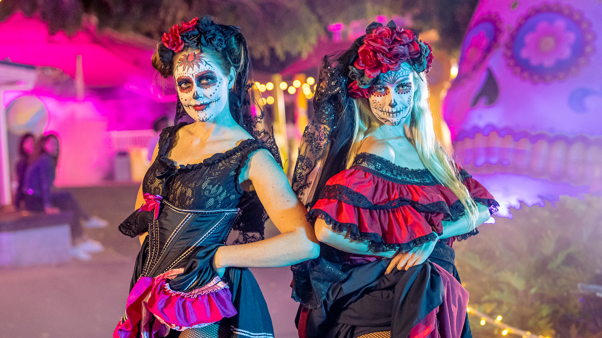 Two women in colorful, ornate Day of the Dead costumes with painted skull faces and flower headpieces pose outdoors at night, with festive lights and decorations in the background.