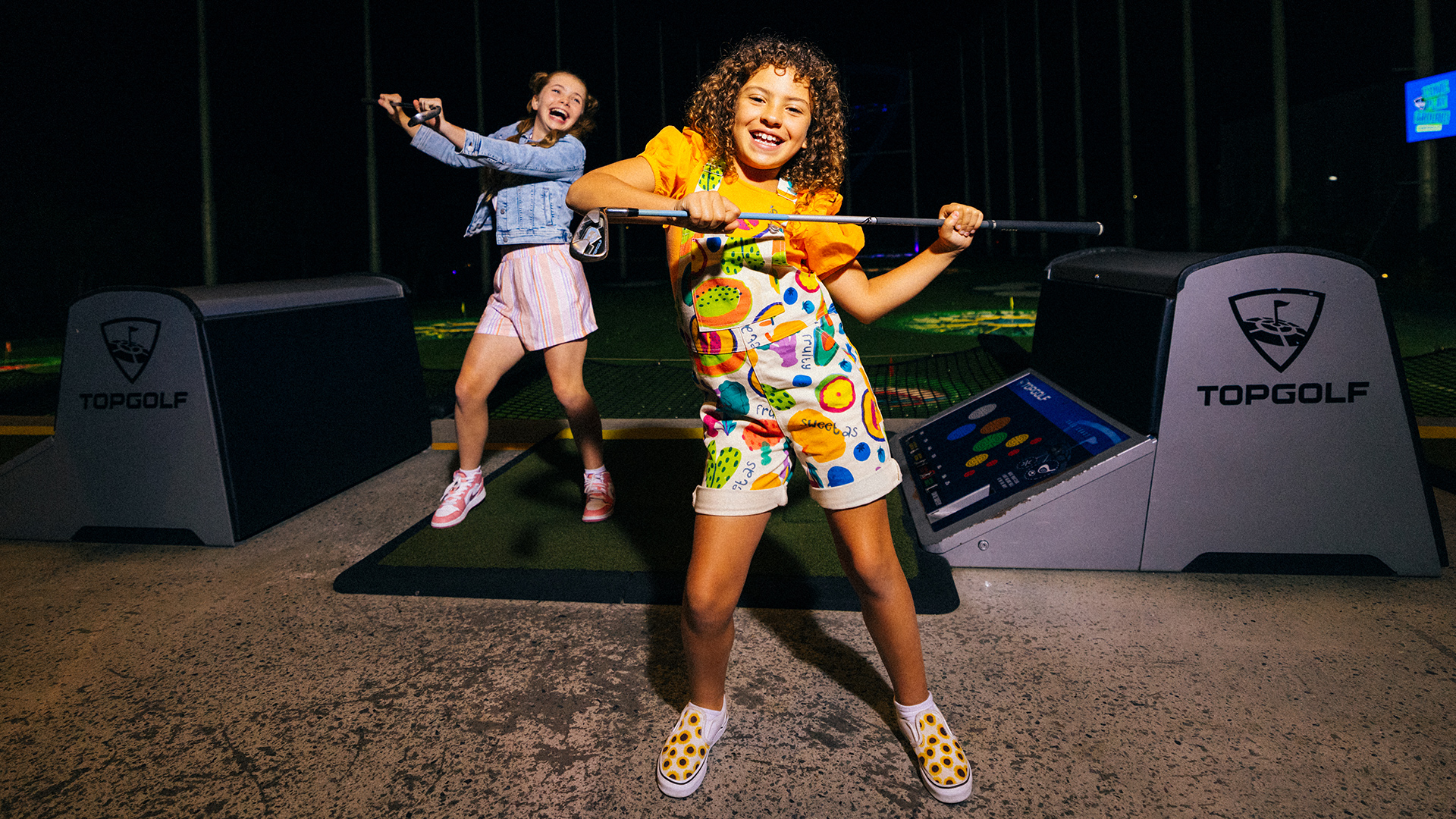 Two smiling girls have fun at a Topgolf location. One girl poses with a golf club in front, while the other stands on the mat behind her, both appearing joyful and energetic in a brightly lit, playful atmosphere.