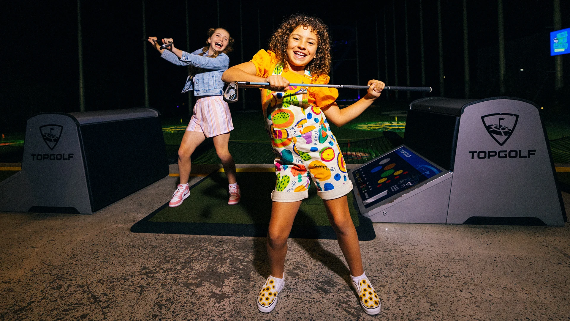 Two smiling girls have fun at a Topgolf location. One girl poses with a golf club in front, while the other stands on the mat behind her, both appearing joyful and energetic in a brightly lit, playful atmosphere.