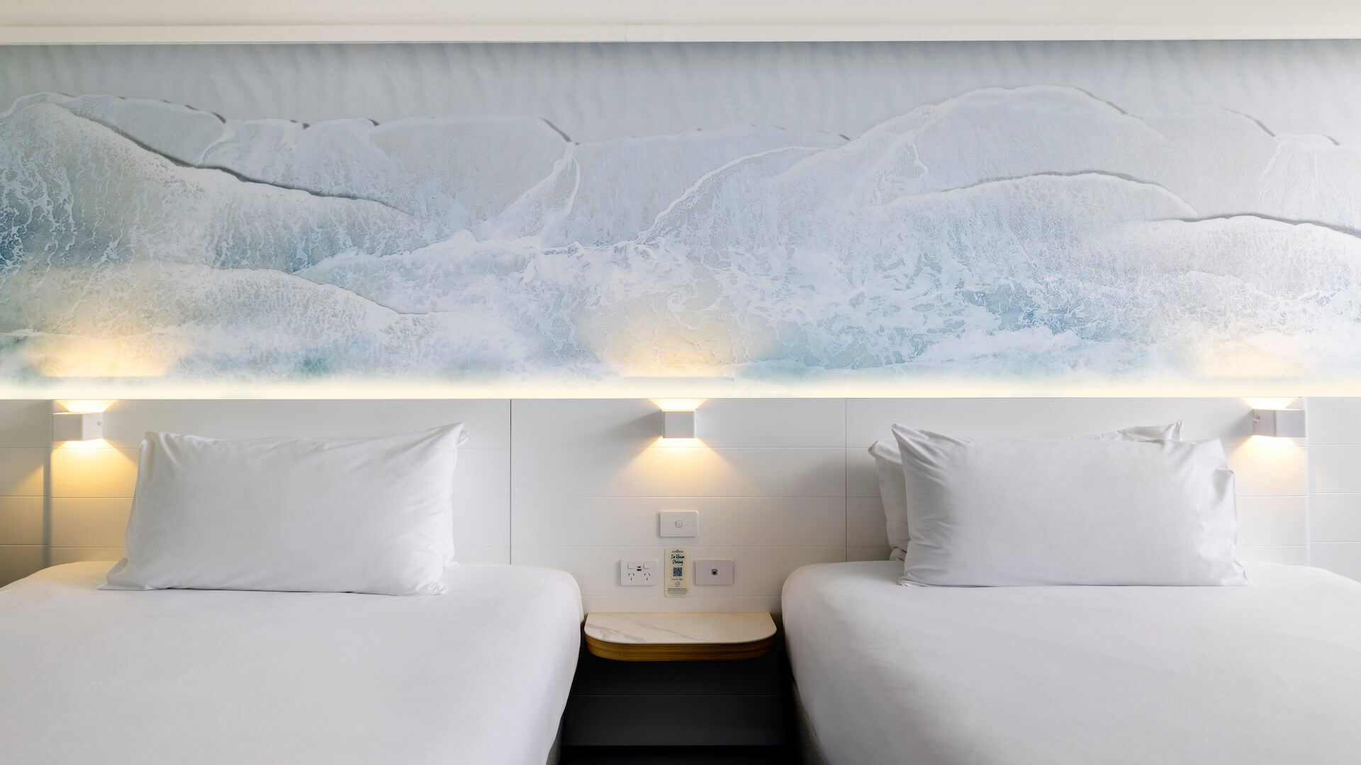 Two neatly made beds with white linens are positioned side by side against a wall featuring a calming ocean wave mural. Soft lighting illuminates the headboard area.