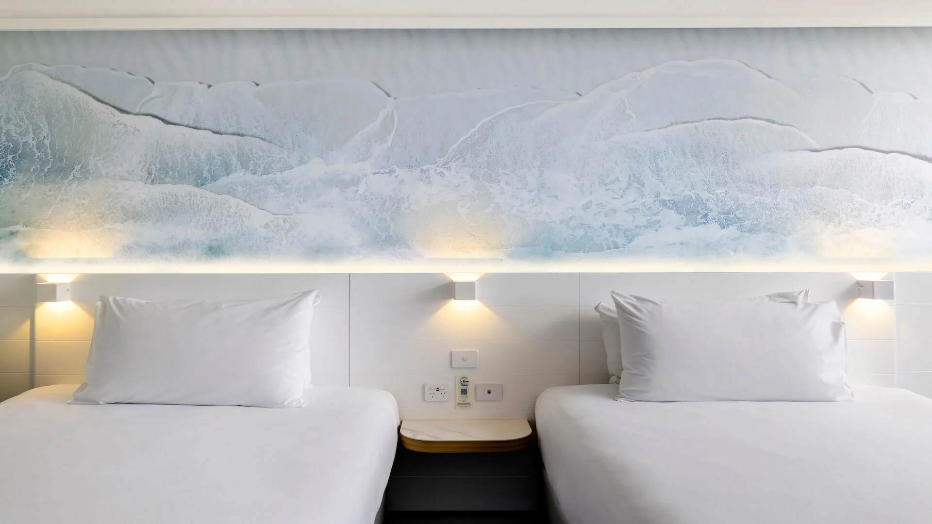 Two neatly made beds with white linens are positioned side by side against a wall featuring a calming ocean wave mural. Soft lighting illuminates the headboard area.
