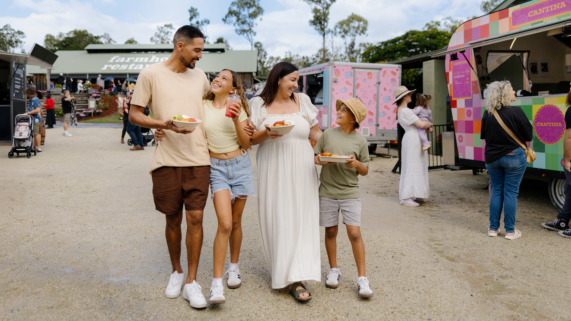 A smiling family of four walks together at an outdoor food market, holding plates of food. Food trucks and other people are visible in the background on a bright day.