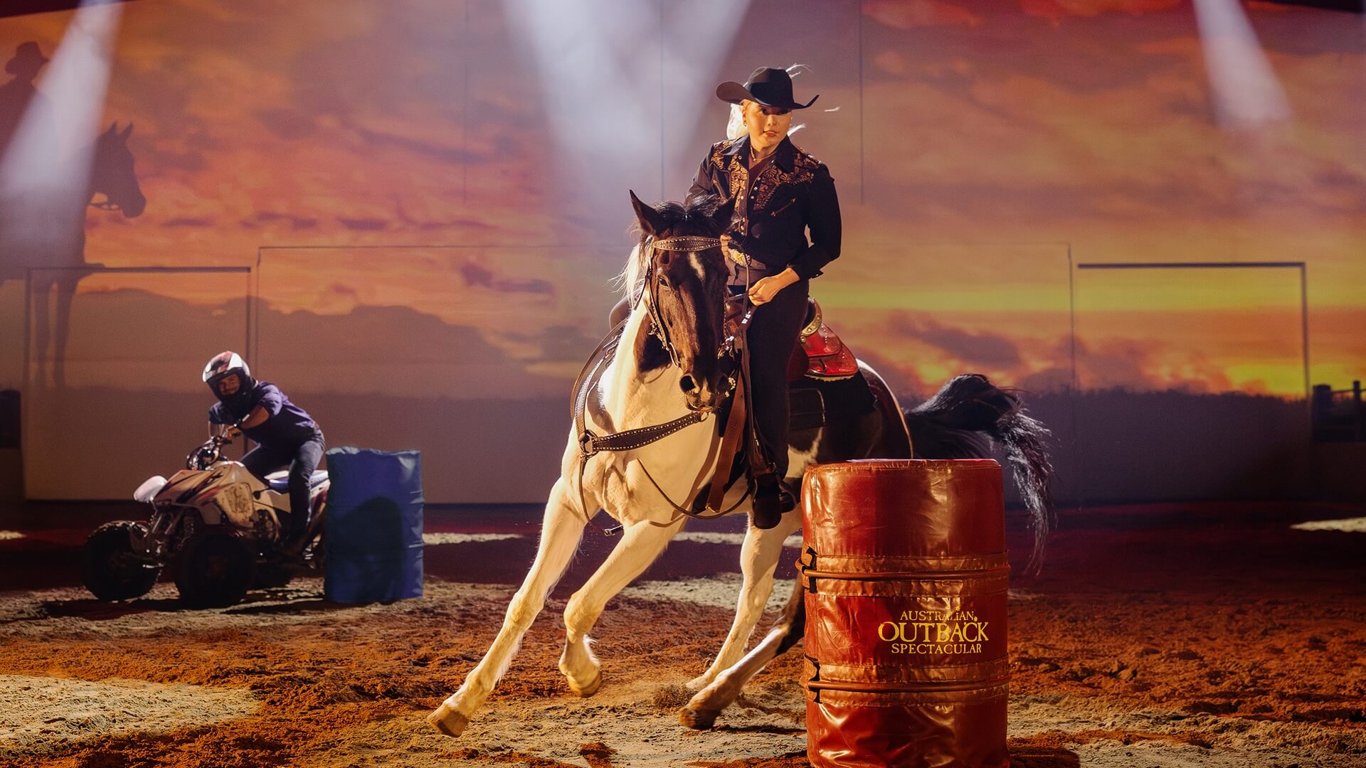 A woman in western attire rides a horse around a barrel in an indoor arena with dramatic stage lighting; a person on an ATV is visible in the background near another barrel.