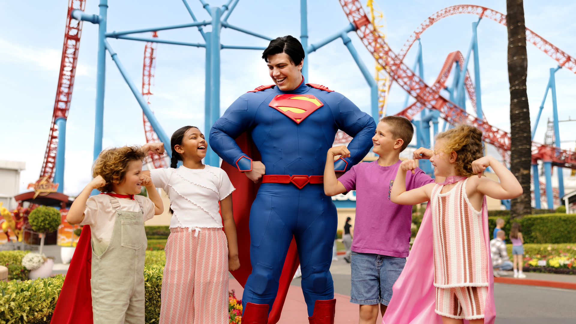 Superman stands smiling with four children wearing capes, all of them flexing their arms and laughing together at an amusement park with roller coasters in the background.