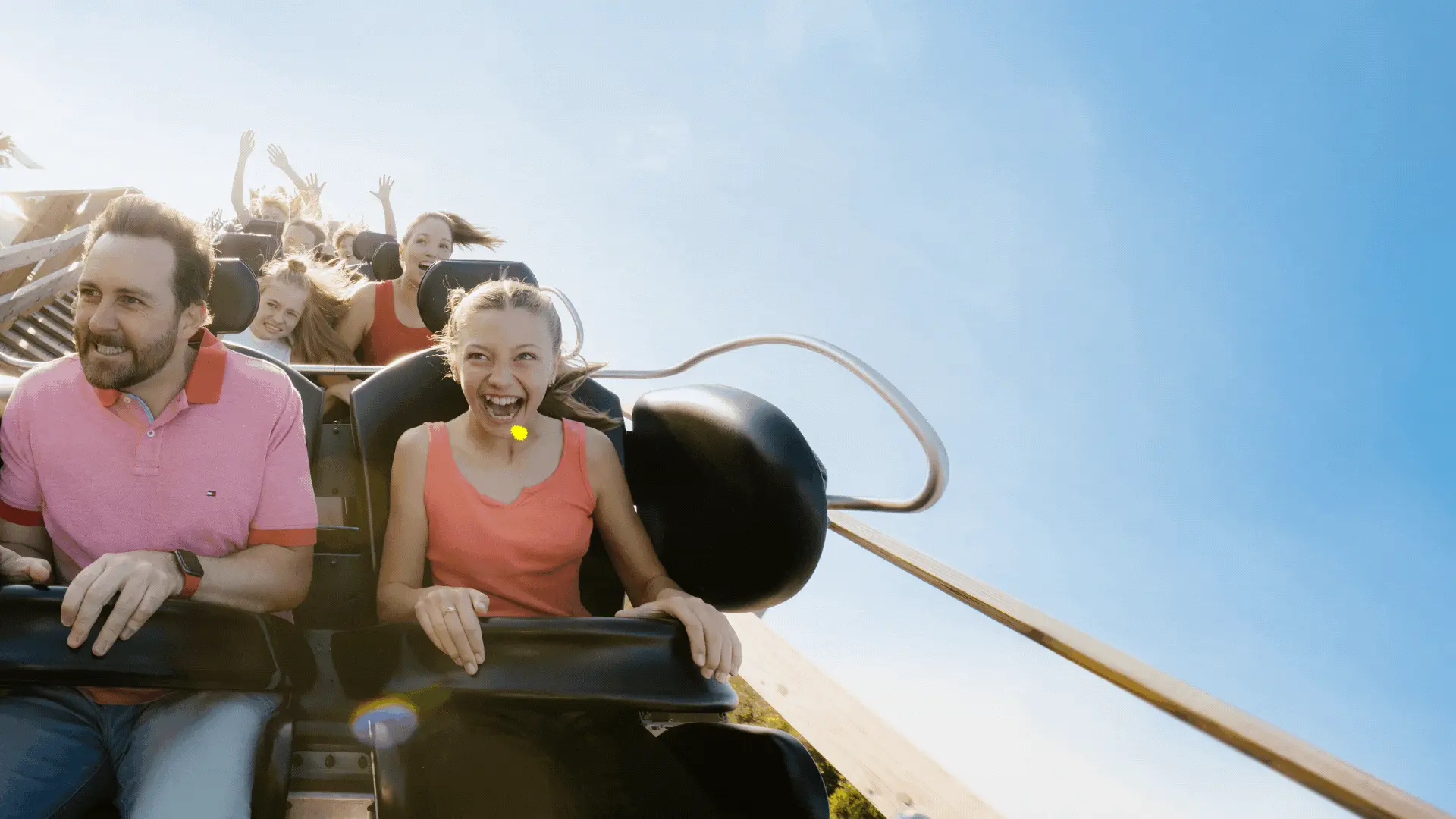 People are riding a roller coaster on a sunny day, laughing and holding on to the safety bars.