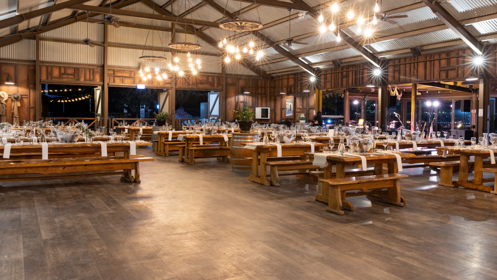 A spacious rustic hall with wooden beams and hanging lights, set up with long wooden tables and benches arranged for an event or banquet. The tables are decorated with glassware and place settings.