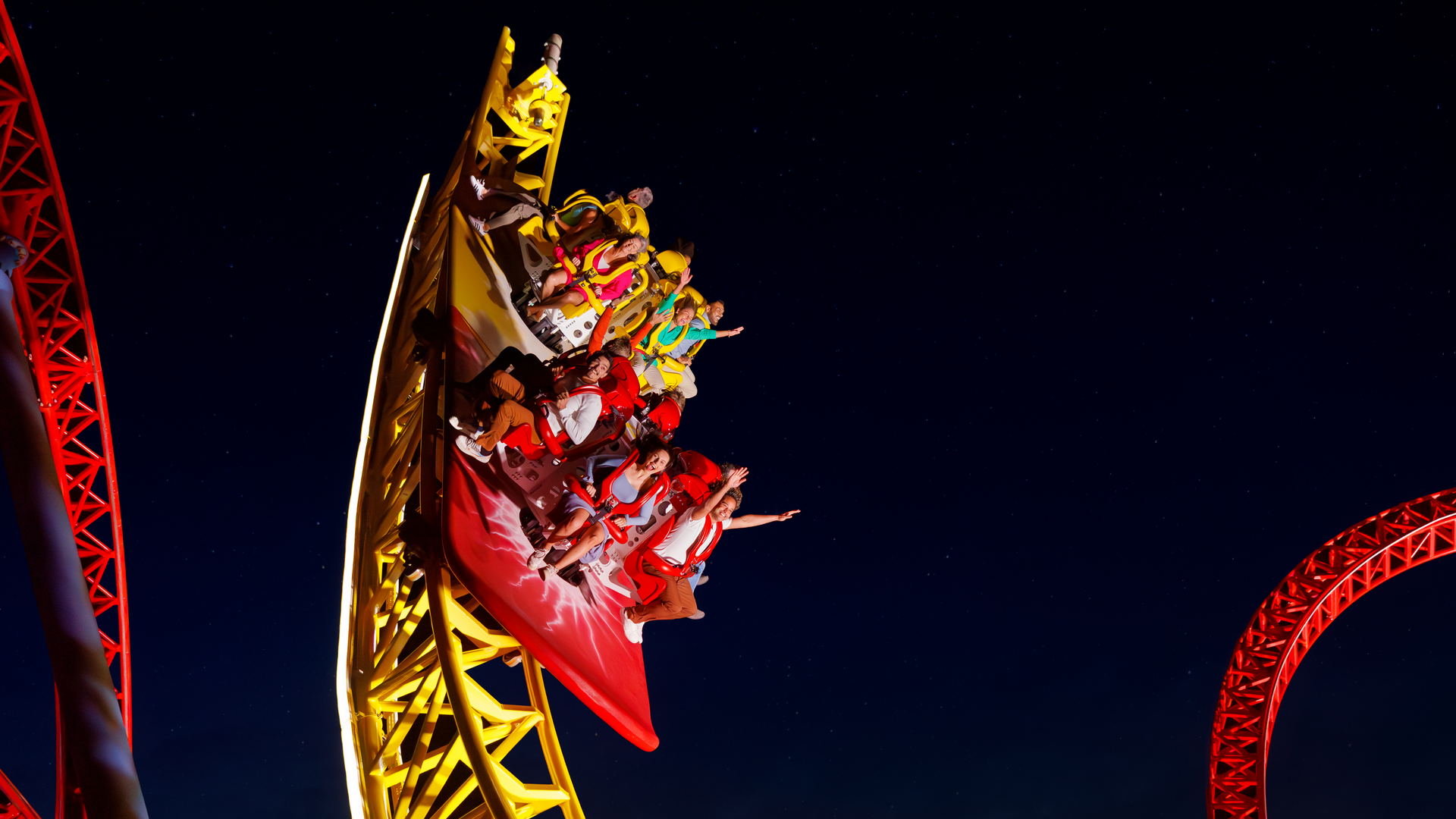 A brightly lit yellow and red roller coaster speeds down a steep curve at night, with riders raising their hands and screaming in excitement against a dark sky.