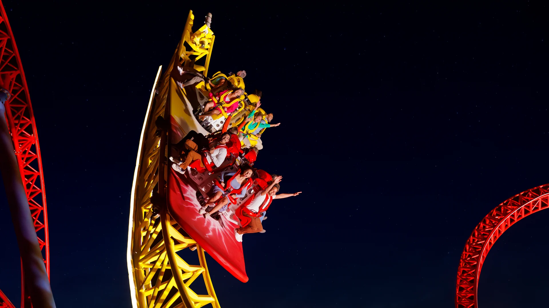 A brightly lit yellow and red roller coaster speeds down a steep curve at night, with riders raising their hands and screaming in excitement against a dark sky.