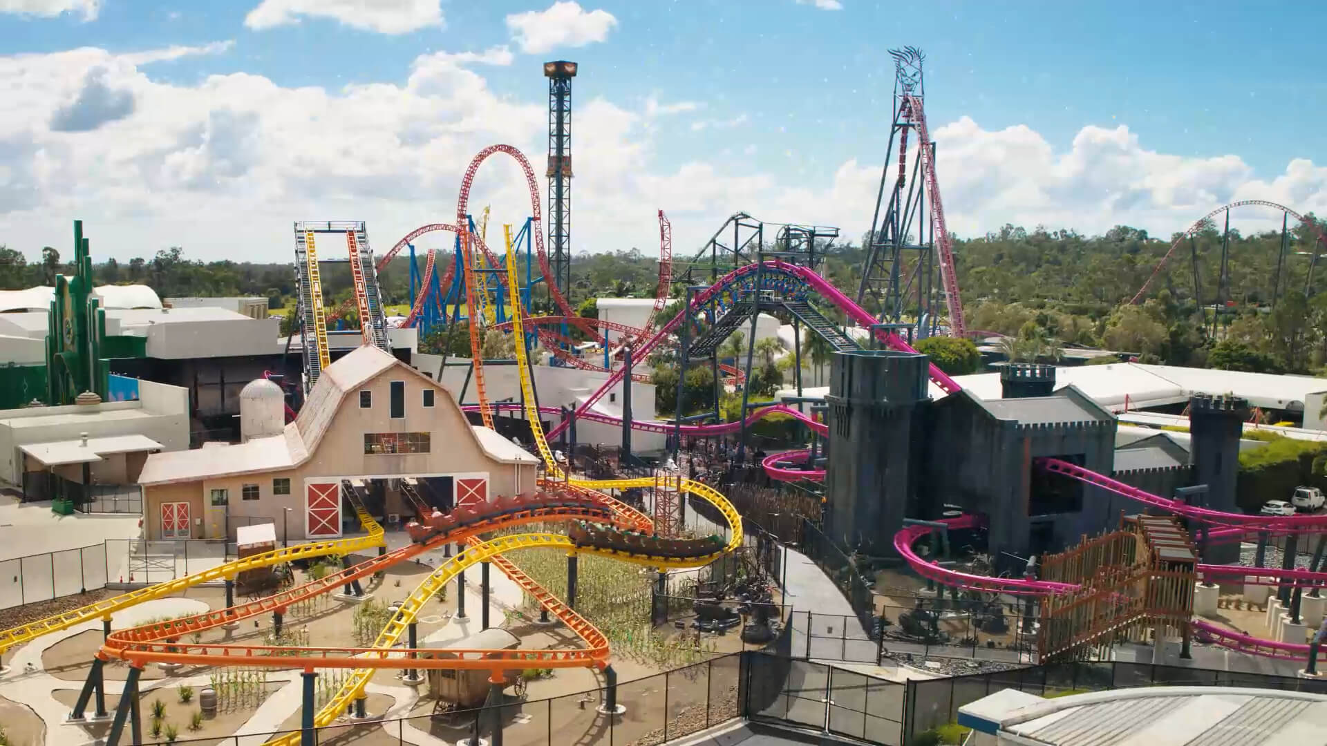 An amusement park with colorful roller coasters, tall rides, and themed buildings surrounded by greenery under a partly cloudy blue sky.