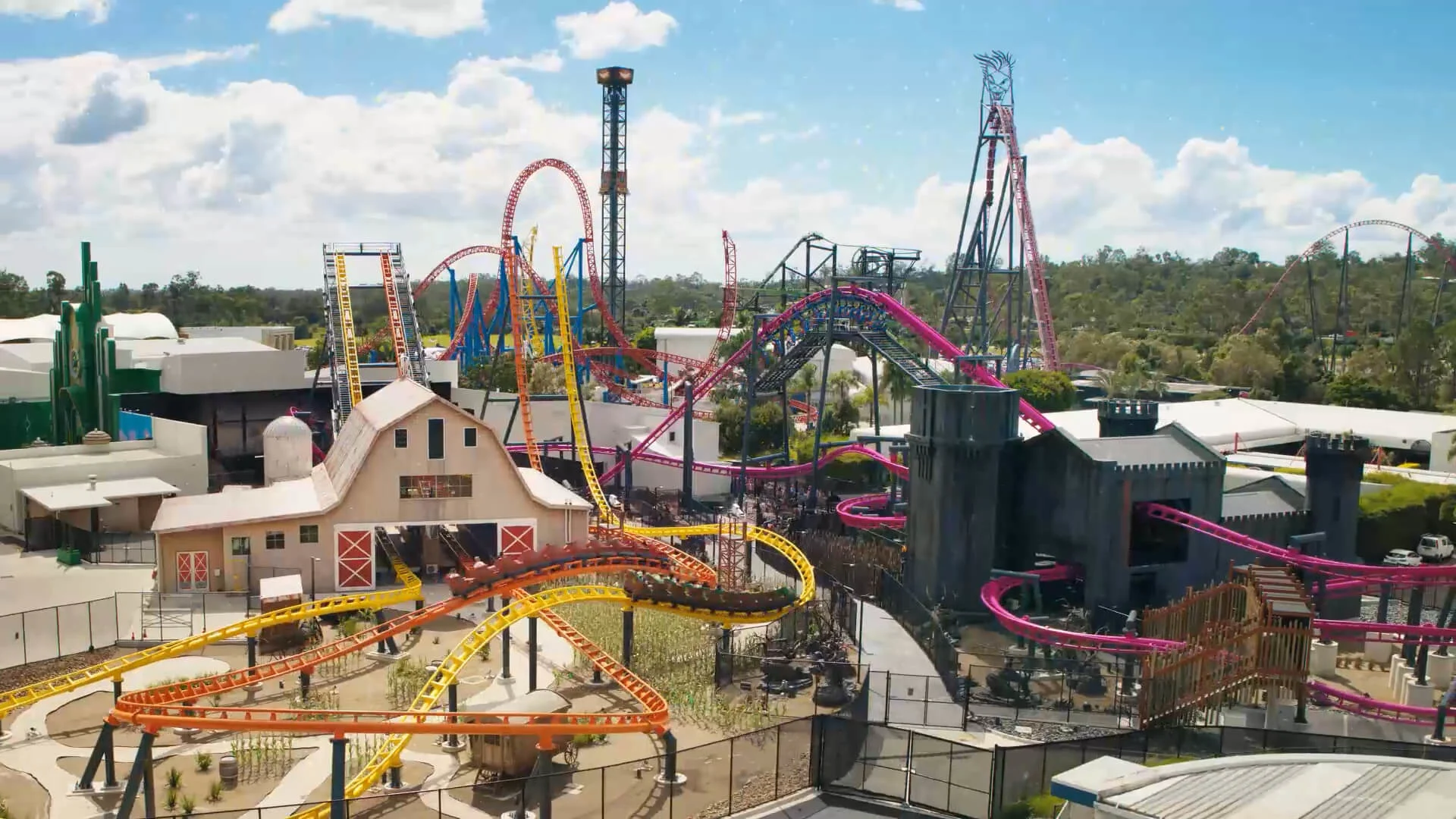 An amusement park with colorful roller coasters, tall rides, and themed buildings surrounded by greenery under a partly cloudy blue sky.