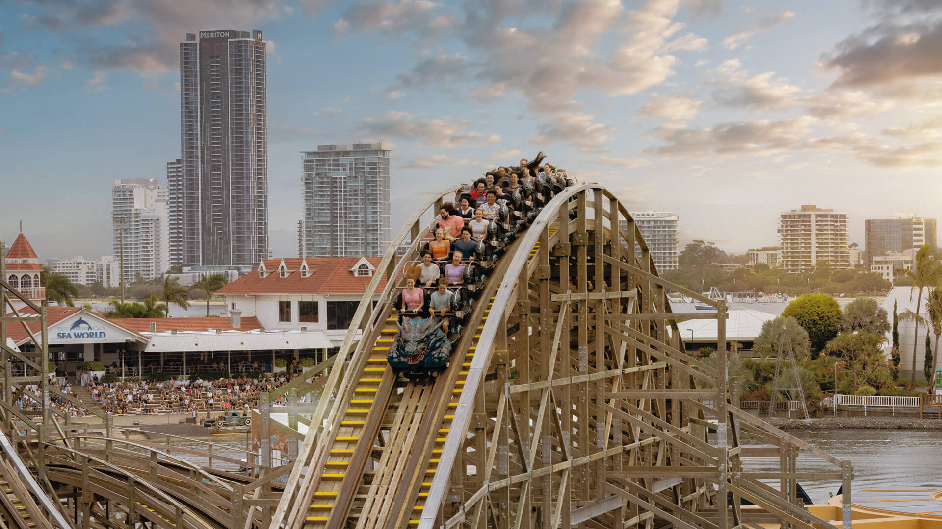 A wooden roller coaster descends at an amusement park, with riders raising their hands; city buildings and a crowd are visible in the background.