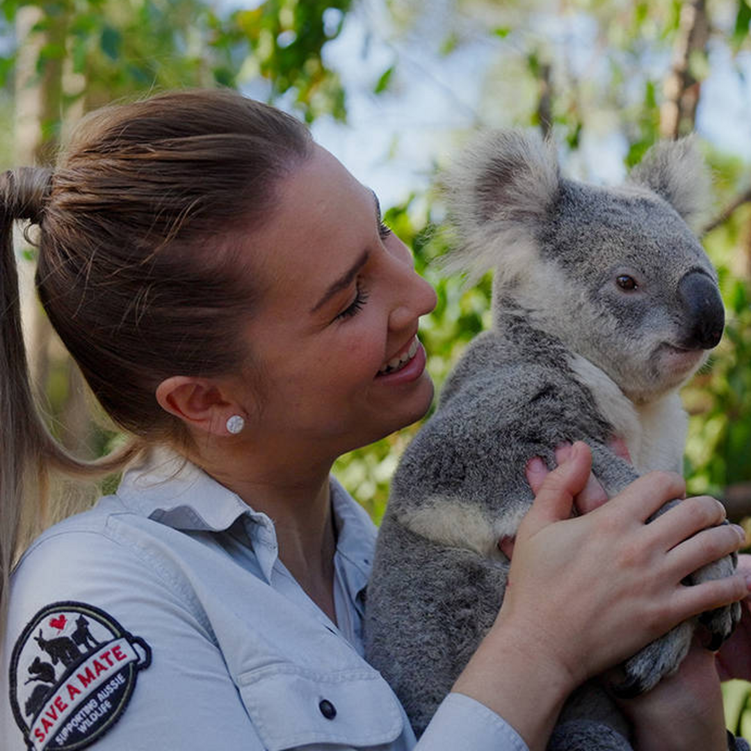 A woman in a light shirt with a "Save a Mate" patch holds a koala while smiling, with green foliage in the background.
