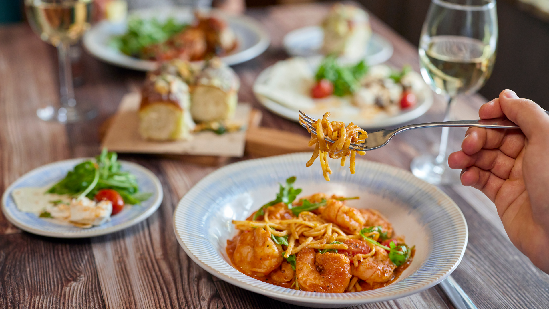 A person holds a fork with spaghetti and shrimp above a plate. On the table are garlic bread, salad, wine, and more plates of food, all set on a wooden surface.