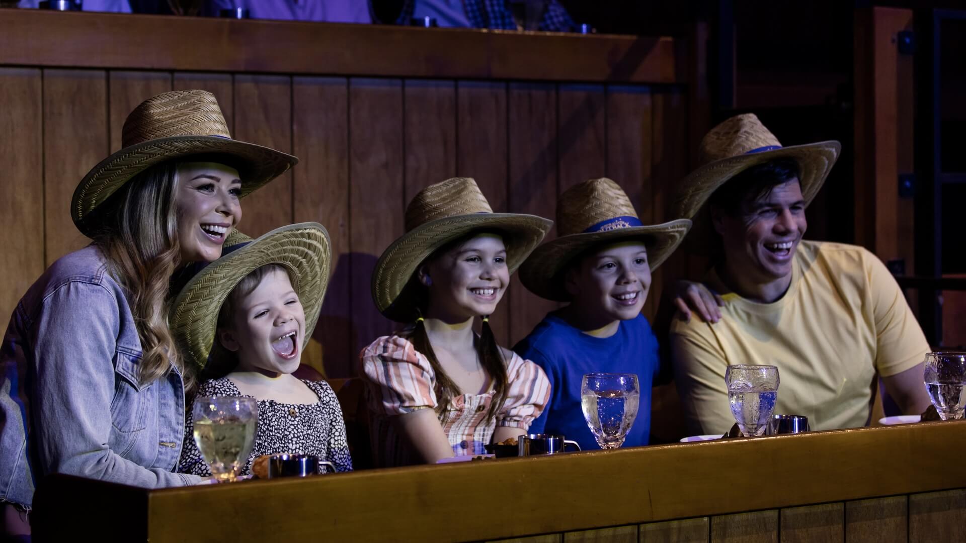 A smiling family of five wearing straw hats sits at a wooden table, watching an event. Three children and two adults are in the front row, with drinks and plates in front of them.