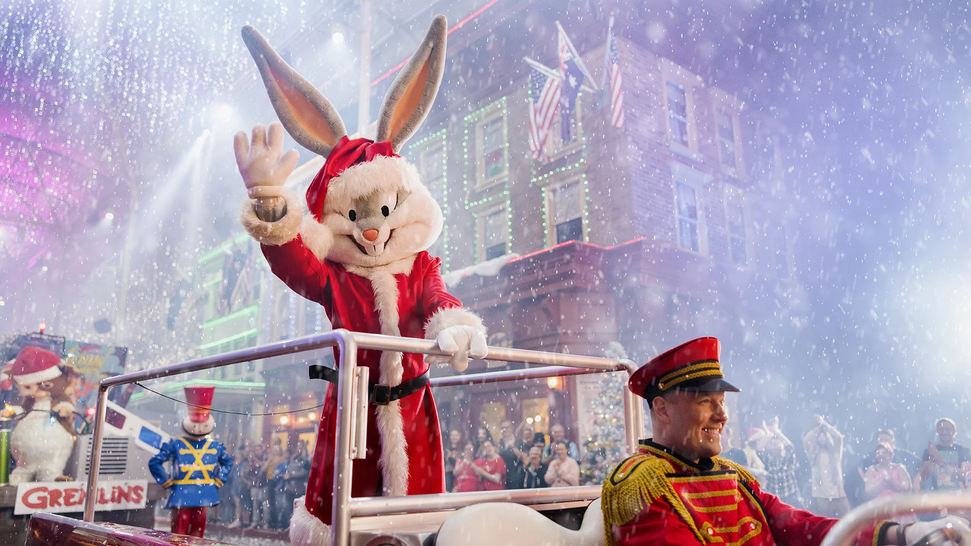 A person in a Bugs Bunny costume dressed as Santa Claus waves from a holiday parade float, surrounded by festive decorations and artificial snow, with a toy soldier and a uniformed man nearby.