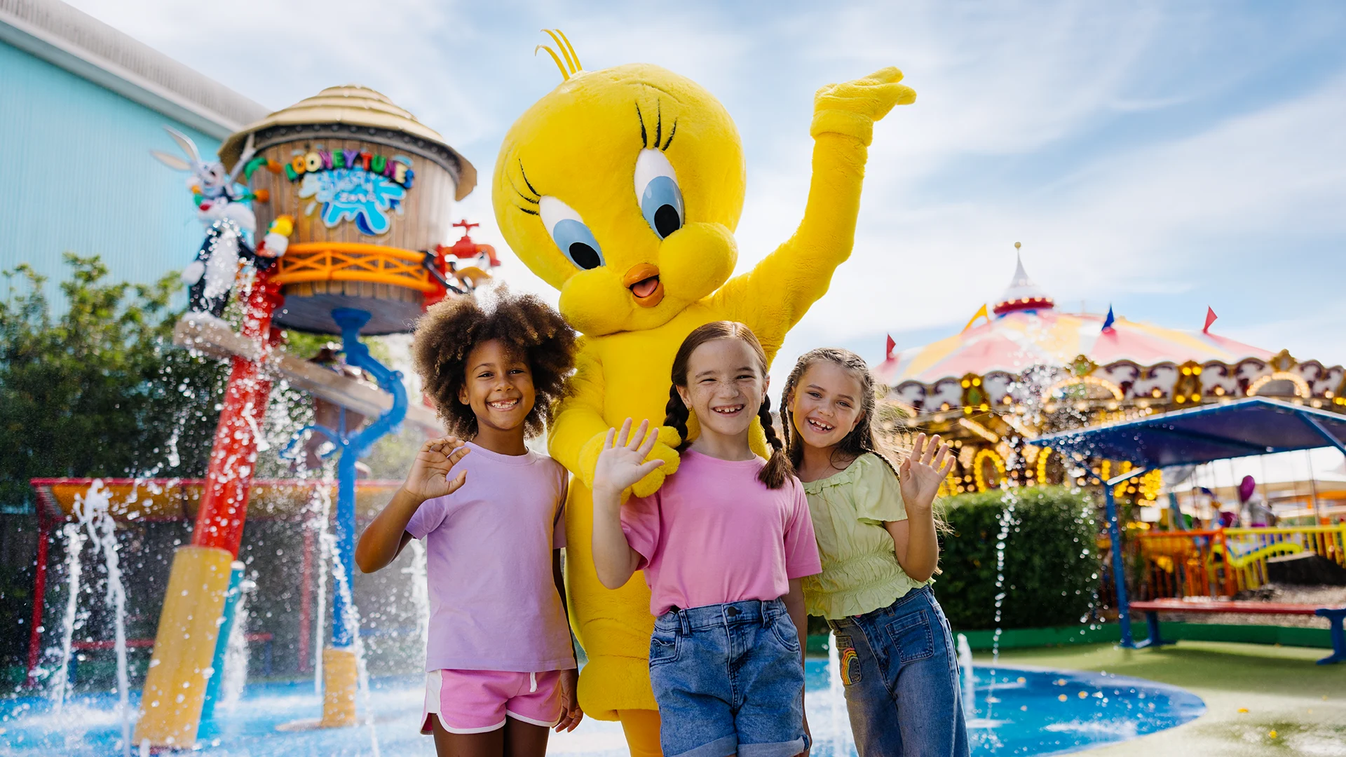 Three smiling children stand in front of a large Tweety Bird mascot, waving by a colorful splash pad and amusement park rides on a sunny day.