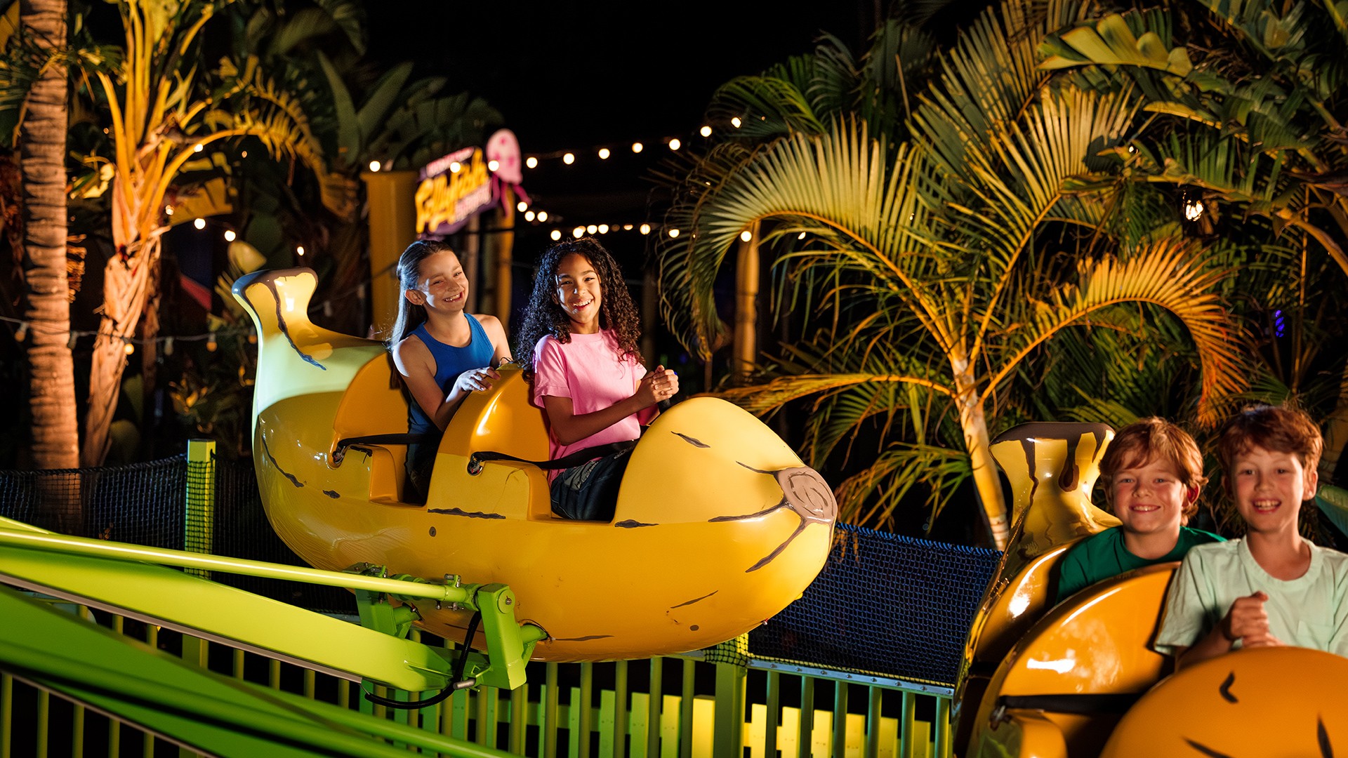Four smiling children ride a bright yellow, banana-shaped amusement park ride at night, surrounded by lush palm trees and festive string lights.
