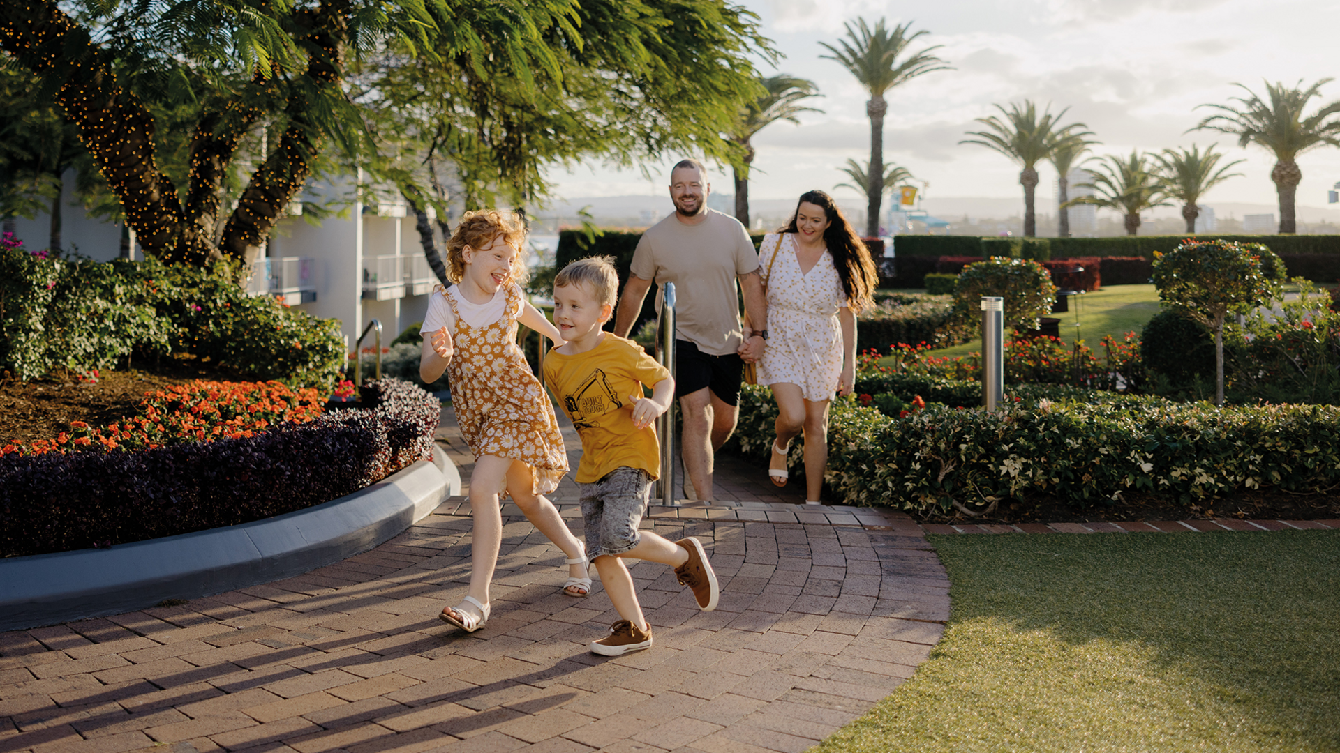 Two children run joyfully along a garden path, followed by two adults under a large tree and surrounded by colorful flowers, greenery, and palm trees on a sunny day.
