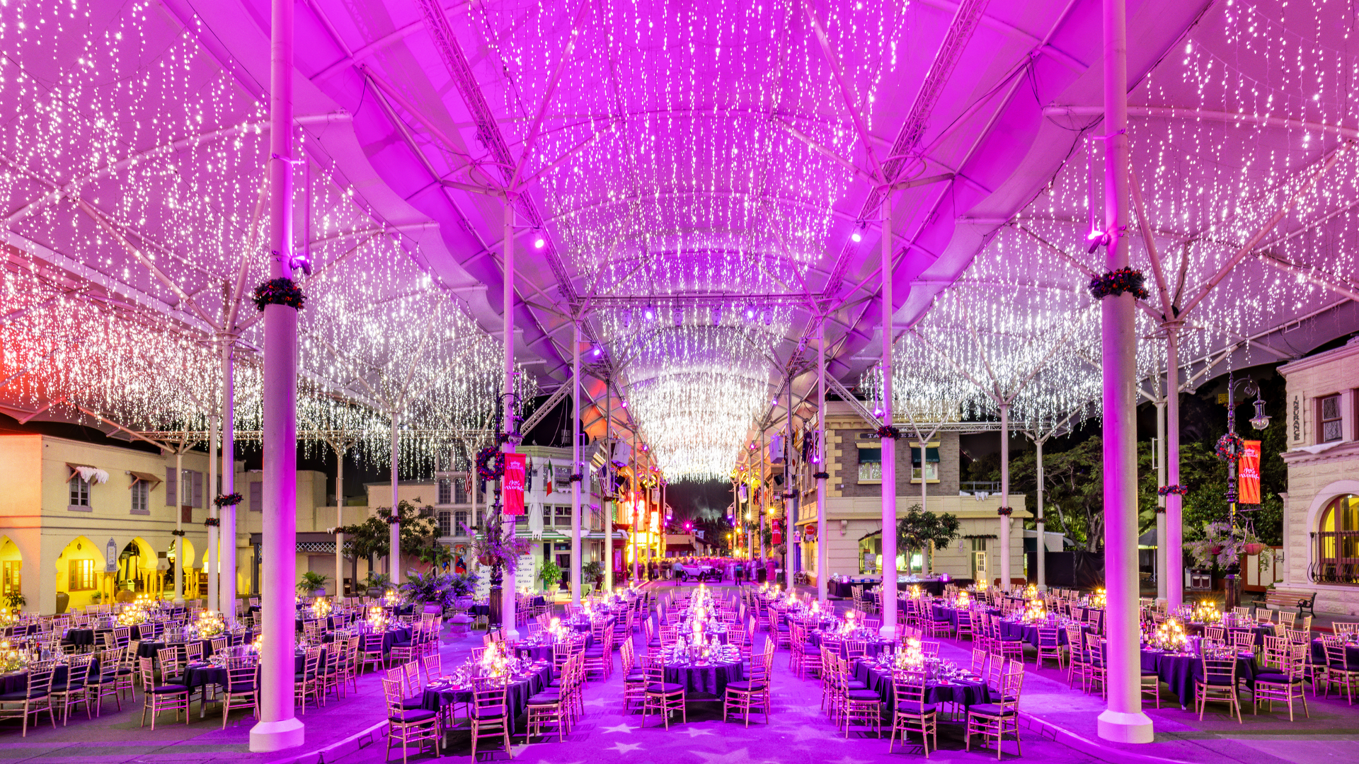 A large outdoor event space is decorated with strings of white fairy lights hanging from the ceiling, casting a purple glow over rows of elegantly set banquet tables and chairs arranged for a festive celebration.
