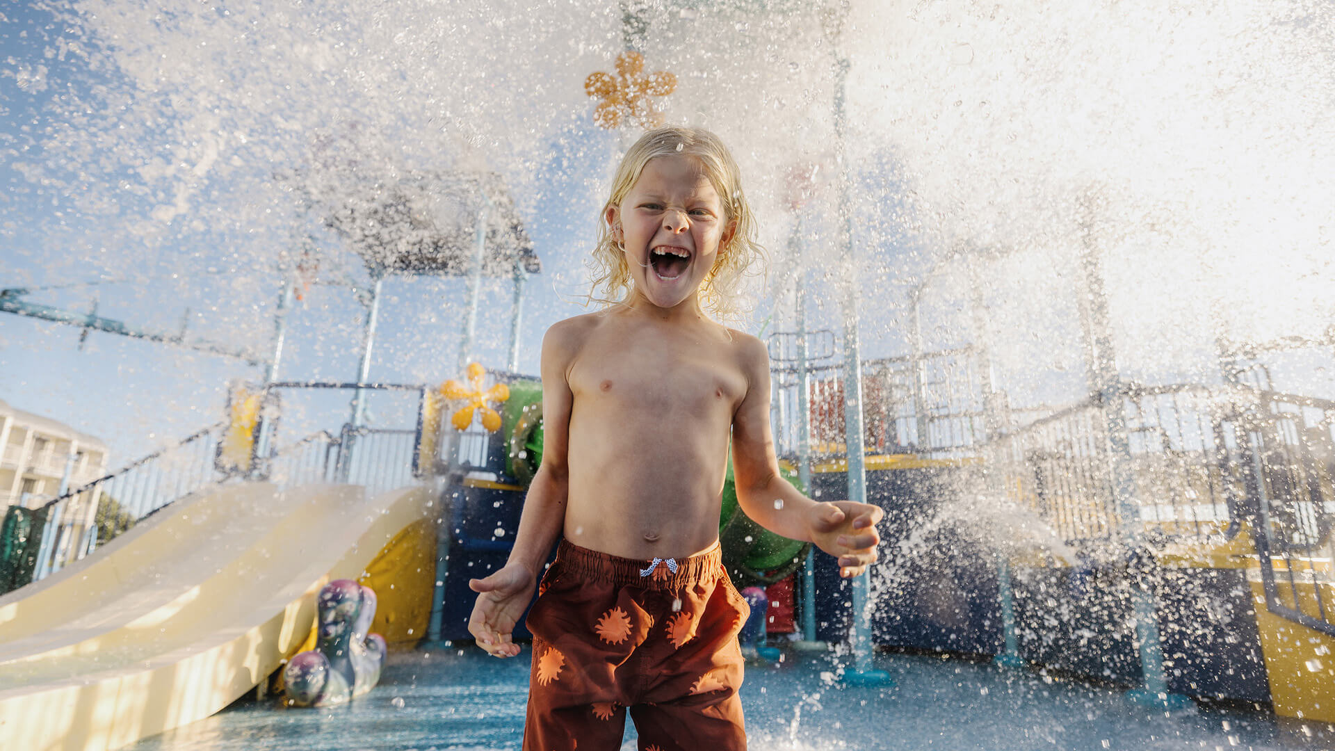 A young child with long blond hair, wearing orange swim trunks, stands laughing joyfully under falling water at an outdoor splash park, with slides and play structures in the background.