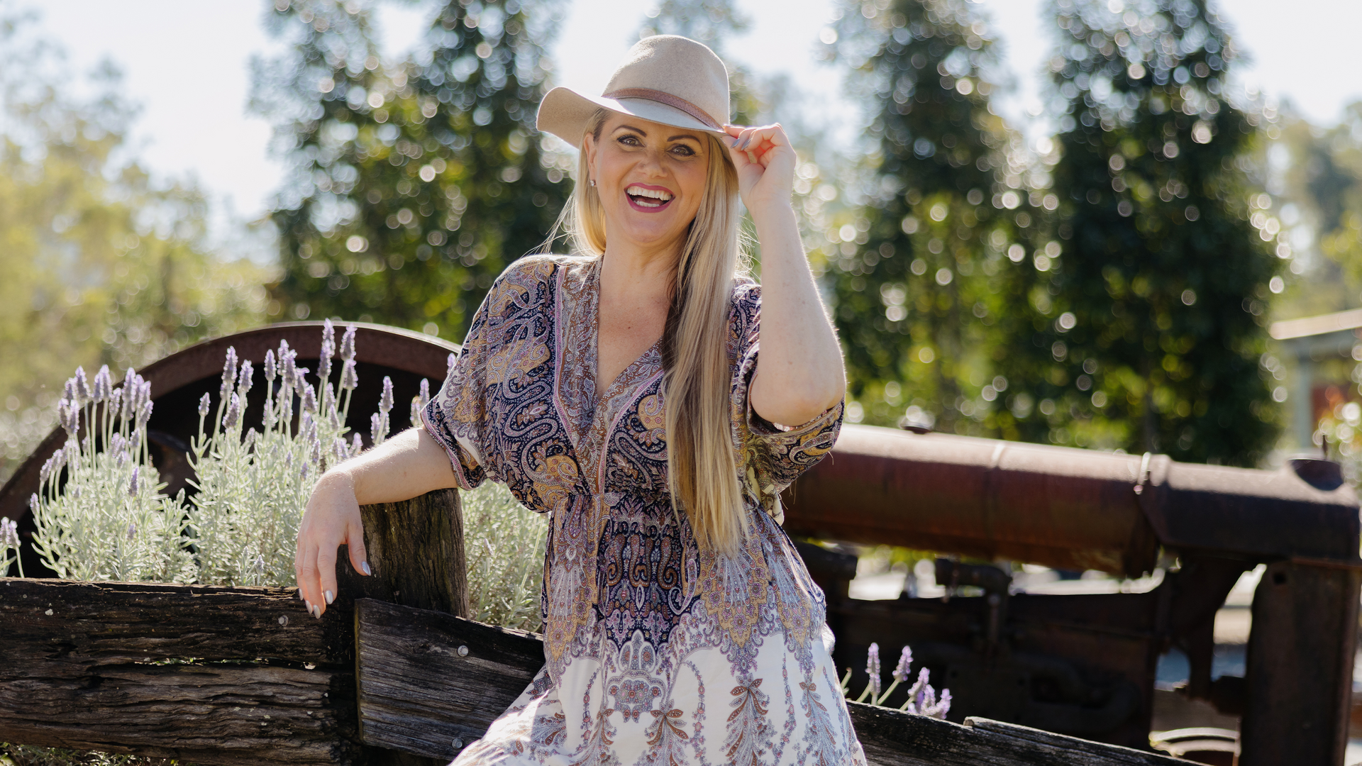 A smiling woman in a patterned dress and wide-brimmed hat sits on a rustic wooden fence surrounded by lavender plants, with trees and an old machine in the background on a sunny day.