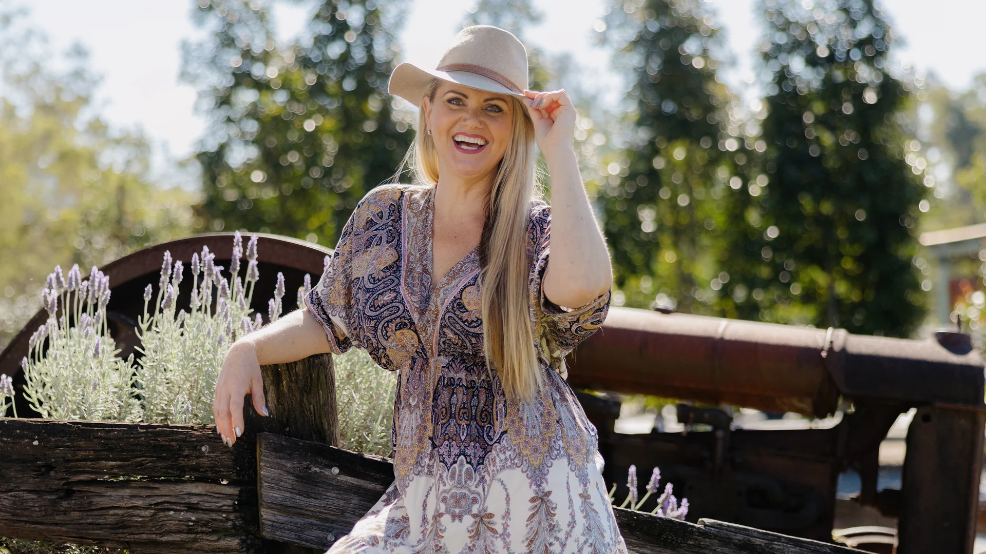A smiling woman in a patterned dress and wide-brimmed hat sits on a rustic wooden fence surrounded by lavender plants, with trees and an old machine in the background on a sunny day.