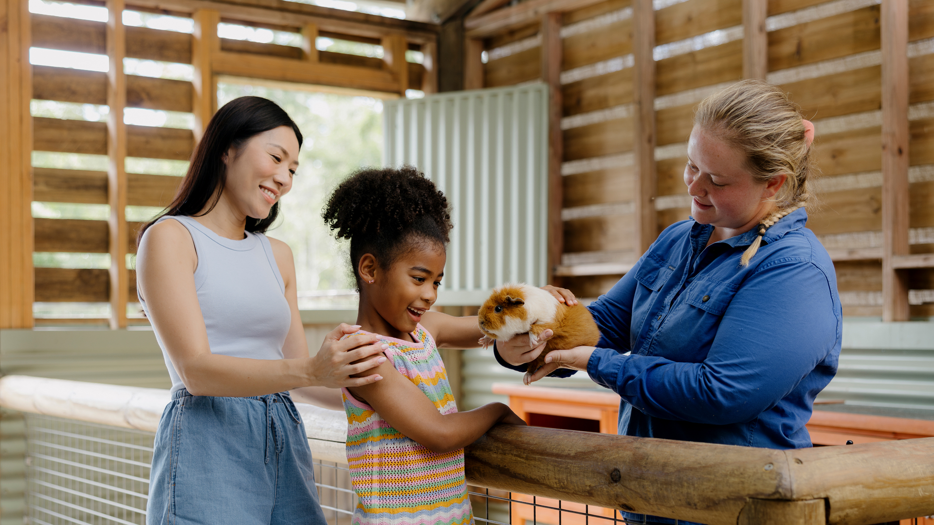 A woman and a young girl smile as another woman holds a guinea pig for them to pet inside a bright, wooden animal enclosure.