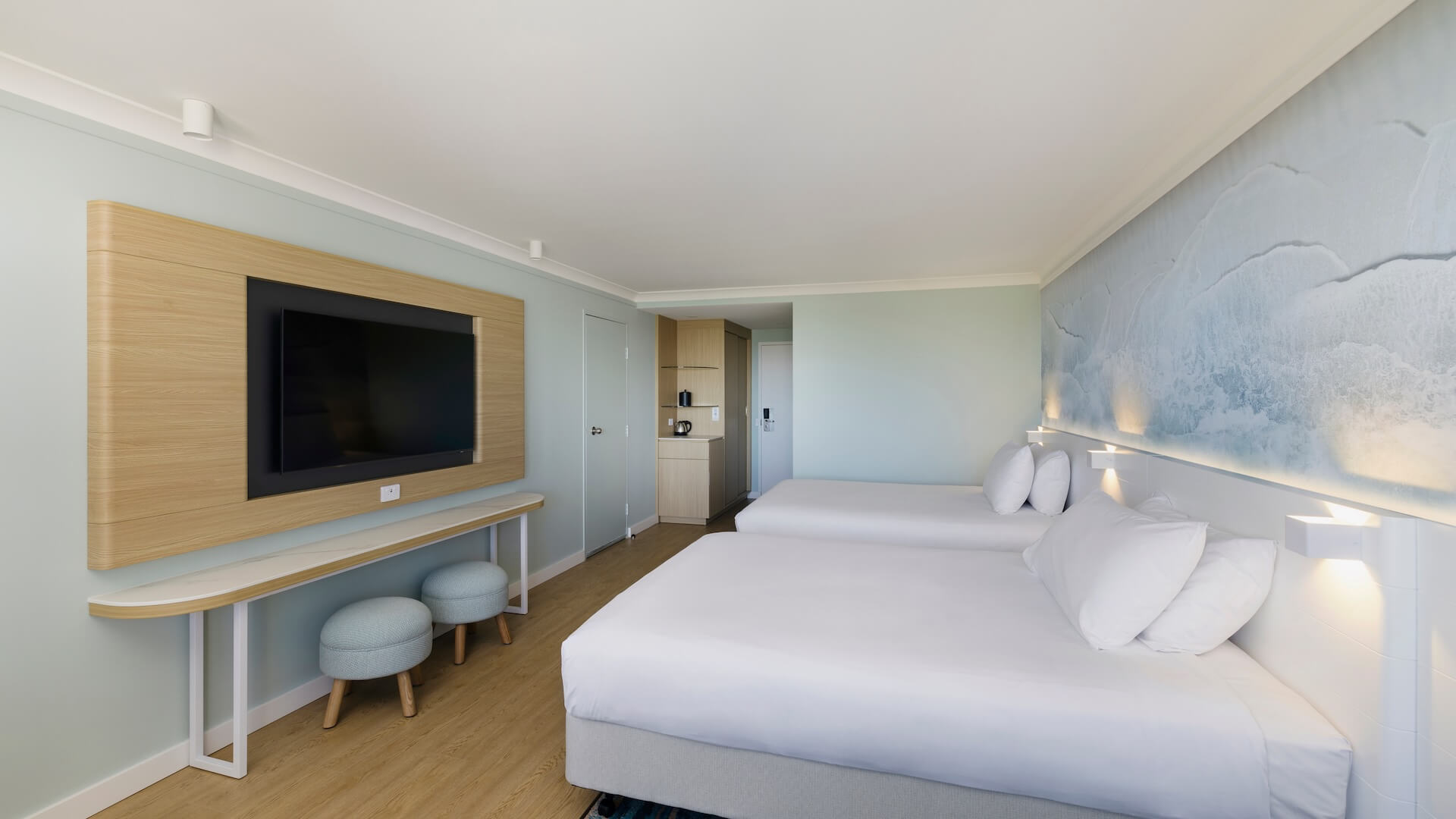 Hotel room with two double beds, a wall-mounted TV, two round stools, wooden flooring, and a wave-themed wall mural.