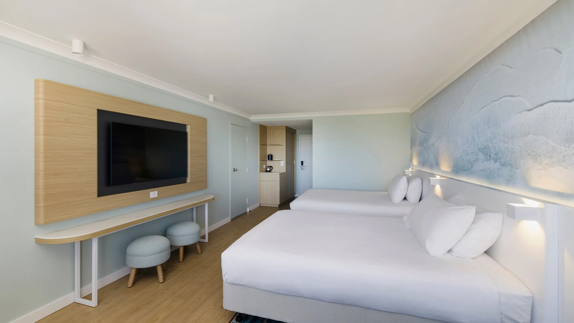 Hotel room with two double beds, a wall-mounted TV, two round stools, wooden flooring, and a wave-themed wall mural.