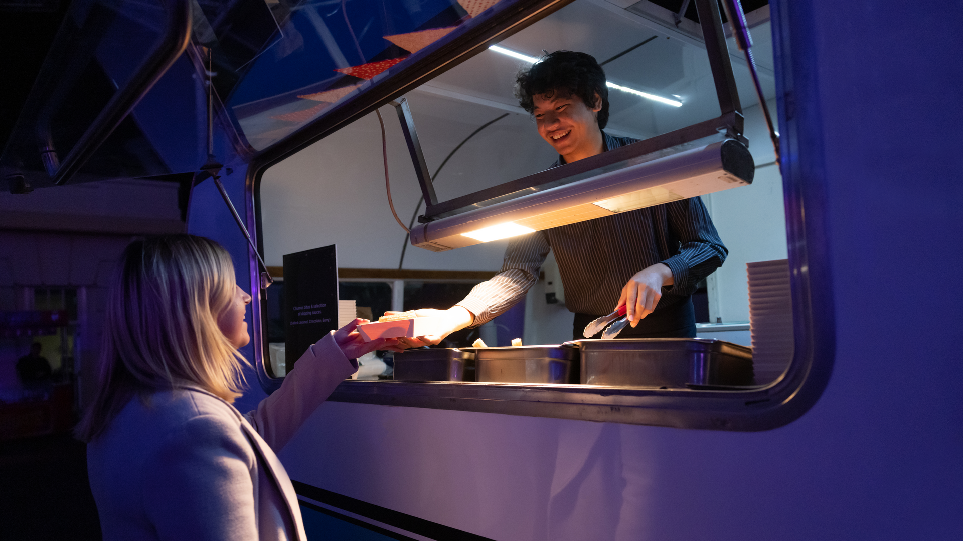 A person in a striped shirt smiles and hands food to a customer through the window of a food truck at night. The customer, wearing a light coat, reaches out to receive the food.