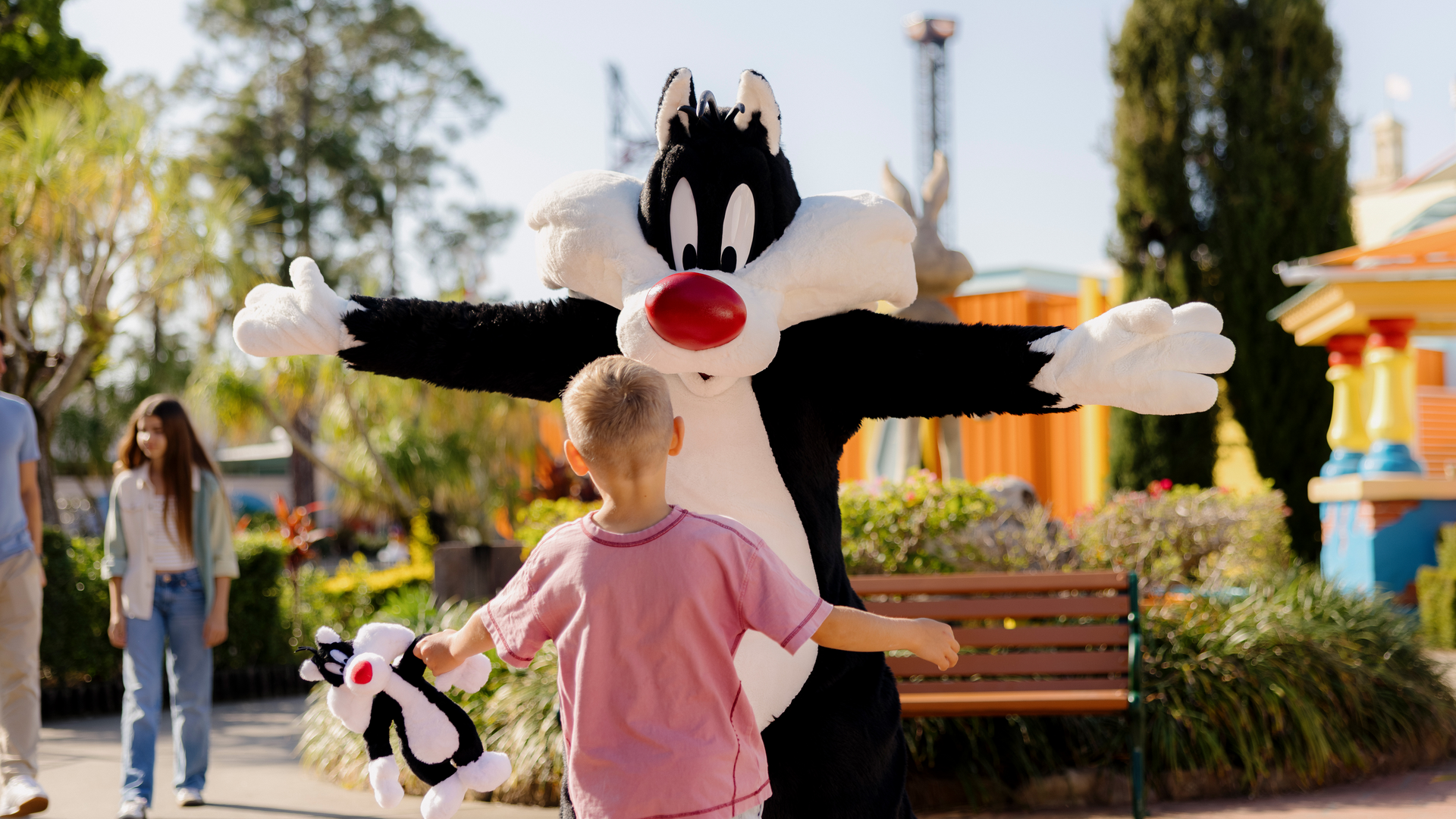 A child holding a small plush toy runs toward a person in a Sylvester the Cat costume with arms open wide in an outdoor park area.