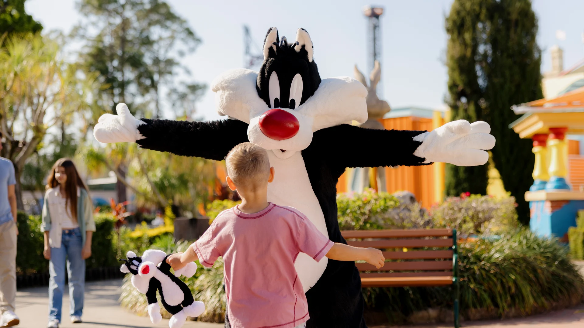 A child holding a small plush toy runs toward a person in a Sylvester the Cat costume with arms open wide in an outdoor park area.