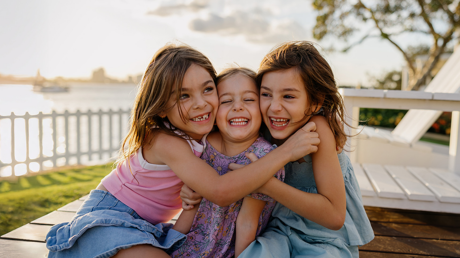 Three young girls sit outdoors by a lakeside, smiling and hugging each other. The sun is setting, creating a warm glow, and a white fence and trees can be seen in the background.