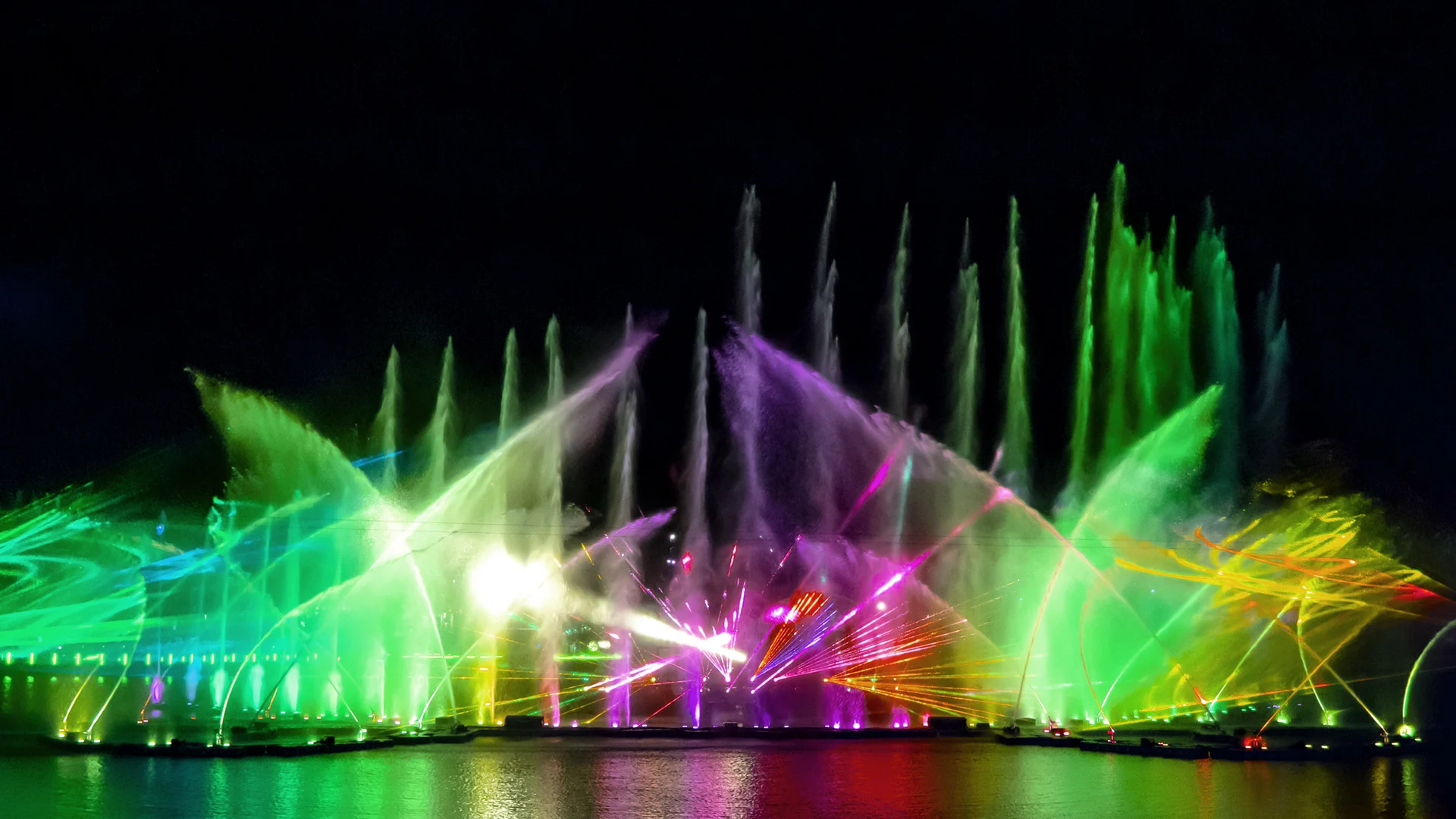 Colorful laser lights and water jets create vibrant patterns over a dark water surface at night, forming a lively and illuminated fountain show with green, yellow, and purple hues.