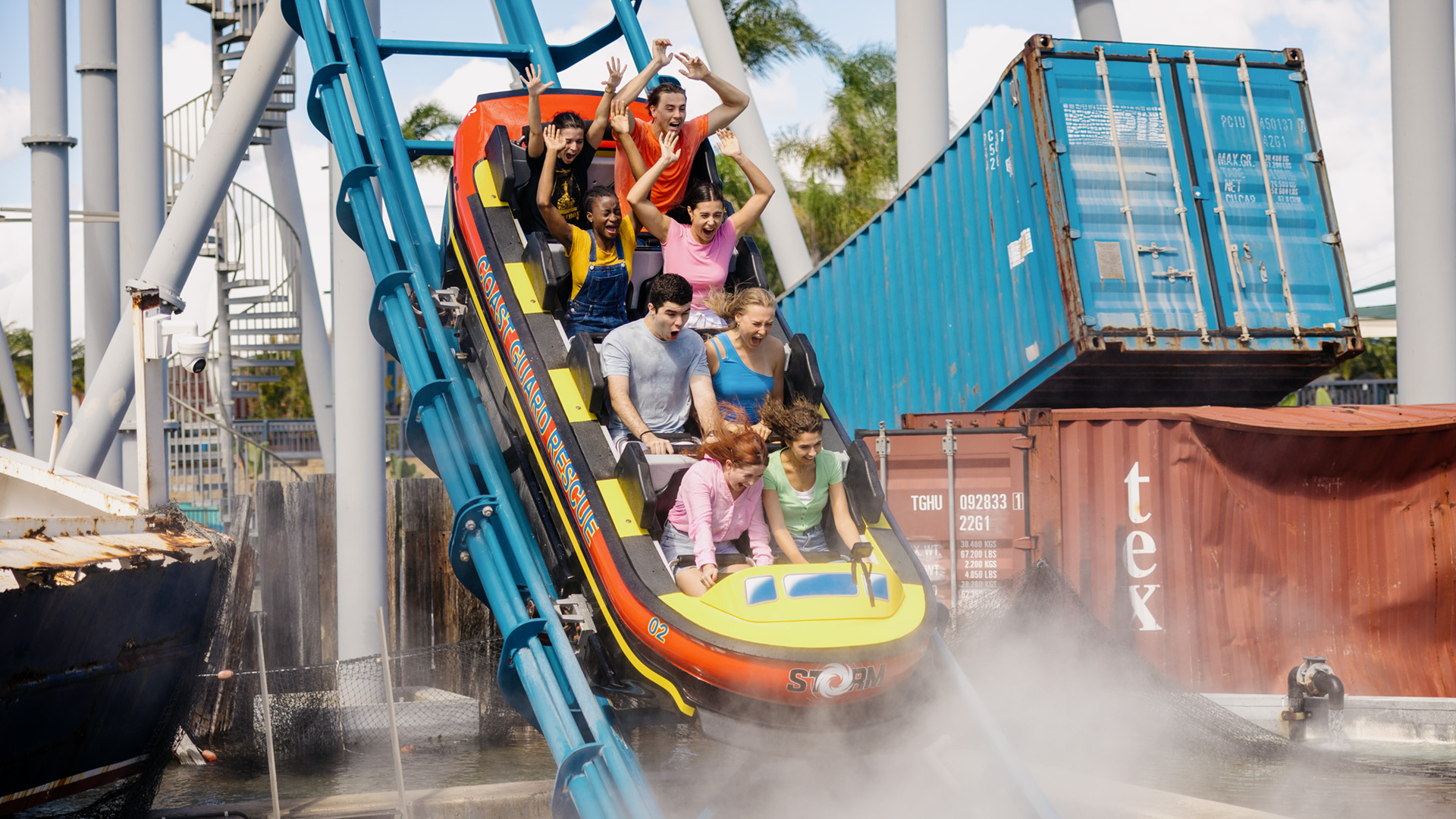 A group of people ride a water coaster down a steep track, splashing through water with shipping containers and steel structures in the background.