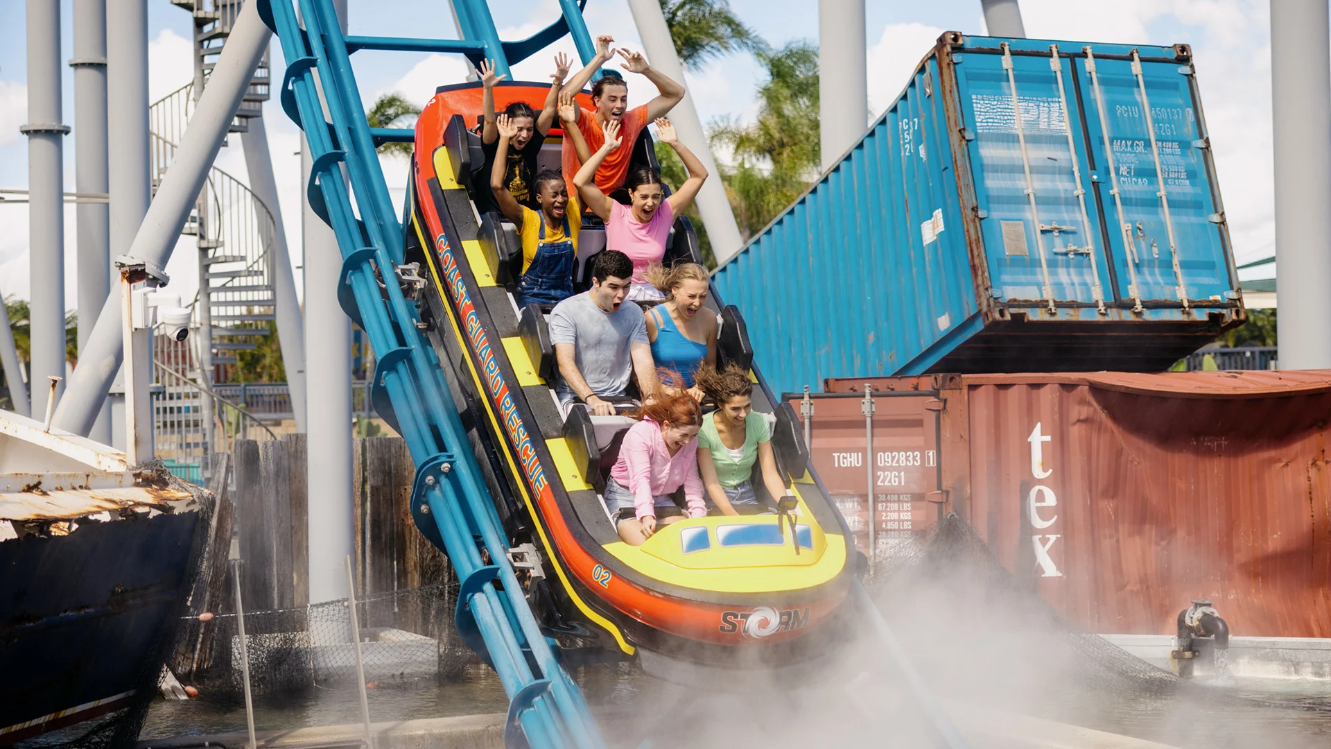 A group of people ride a water coaster down a steep track, splashing through water with shipping containers and steel structures in the background.