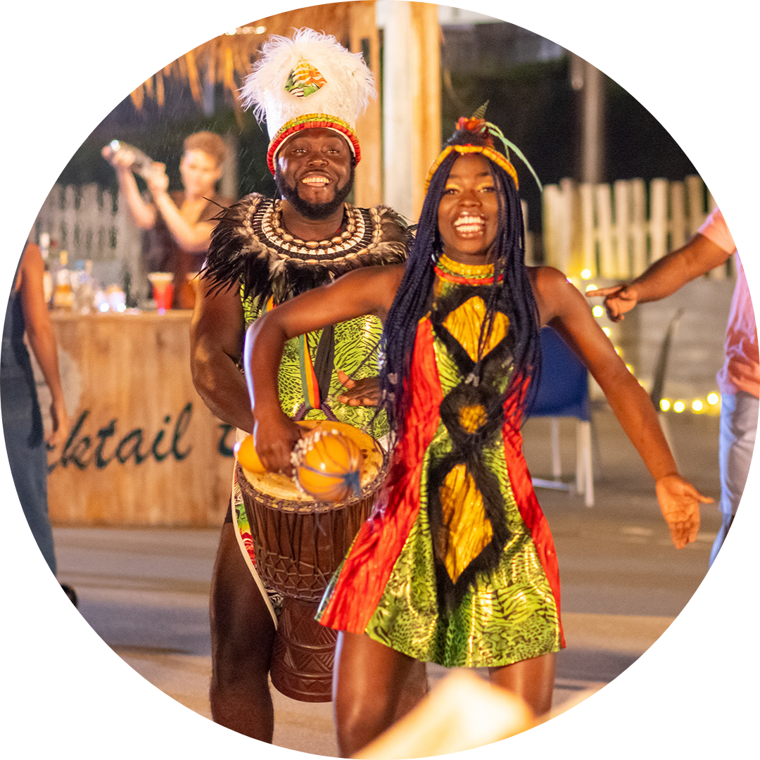 Two people in colorful traditional costumes dance and play a drum at an outdoor event, smiling and celebrating. There are lights and people in the background near a wooden bar.