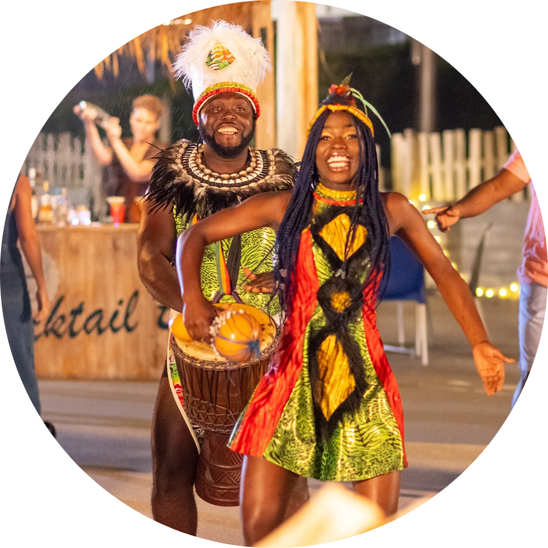 Two people in colorful traditional costumes dance and play a drum at an outdoor event, smiling and celebrating. There are lights and people in the background near a wooden bar.