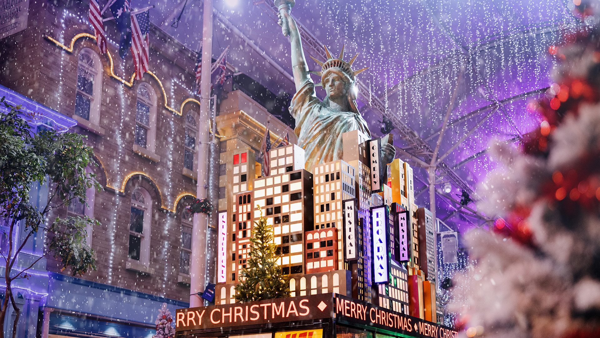 A festive city scene features a replica of the Statue of Liberty surrounded by colorful buildings, holiday lights, falling snow, and a sign that reads “Merry Christmas.”.