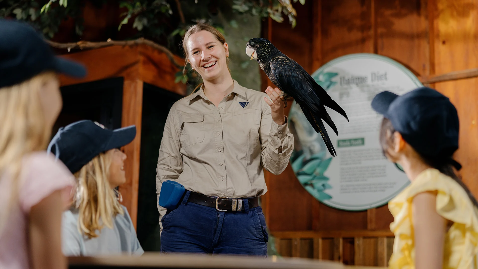 A smiling woman in a khaki shirt holds a black parrot while talking to three children in caps, who are seated and attentively watching her. They are indoors, with a wooden wall and a sign in the background.