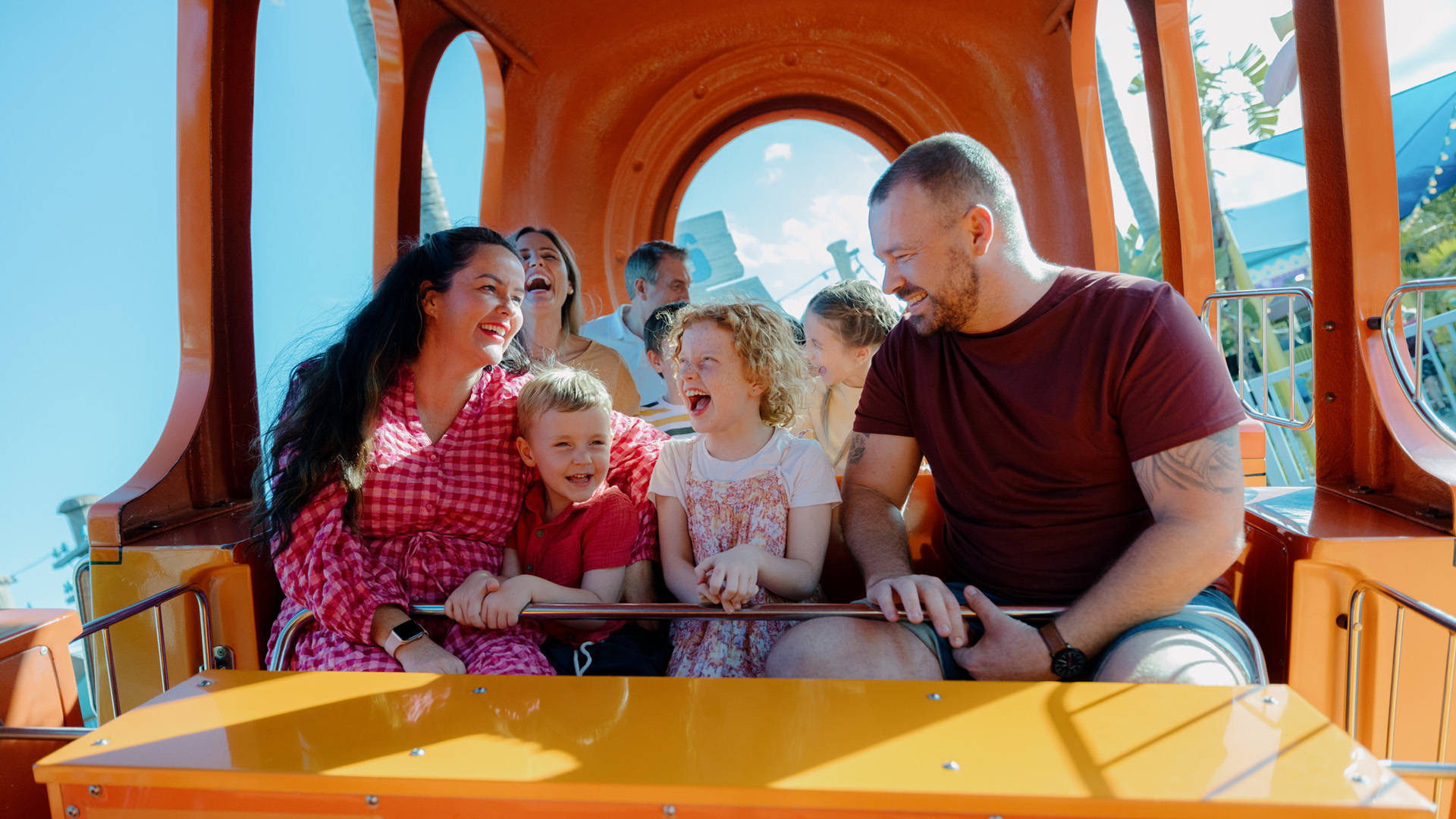A family of six enjoys a ride inside an orange amusement park cabin, smiling and laughing under a clear blue sky.