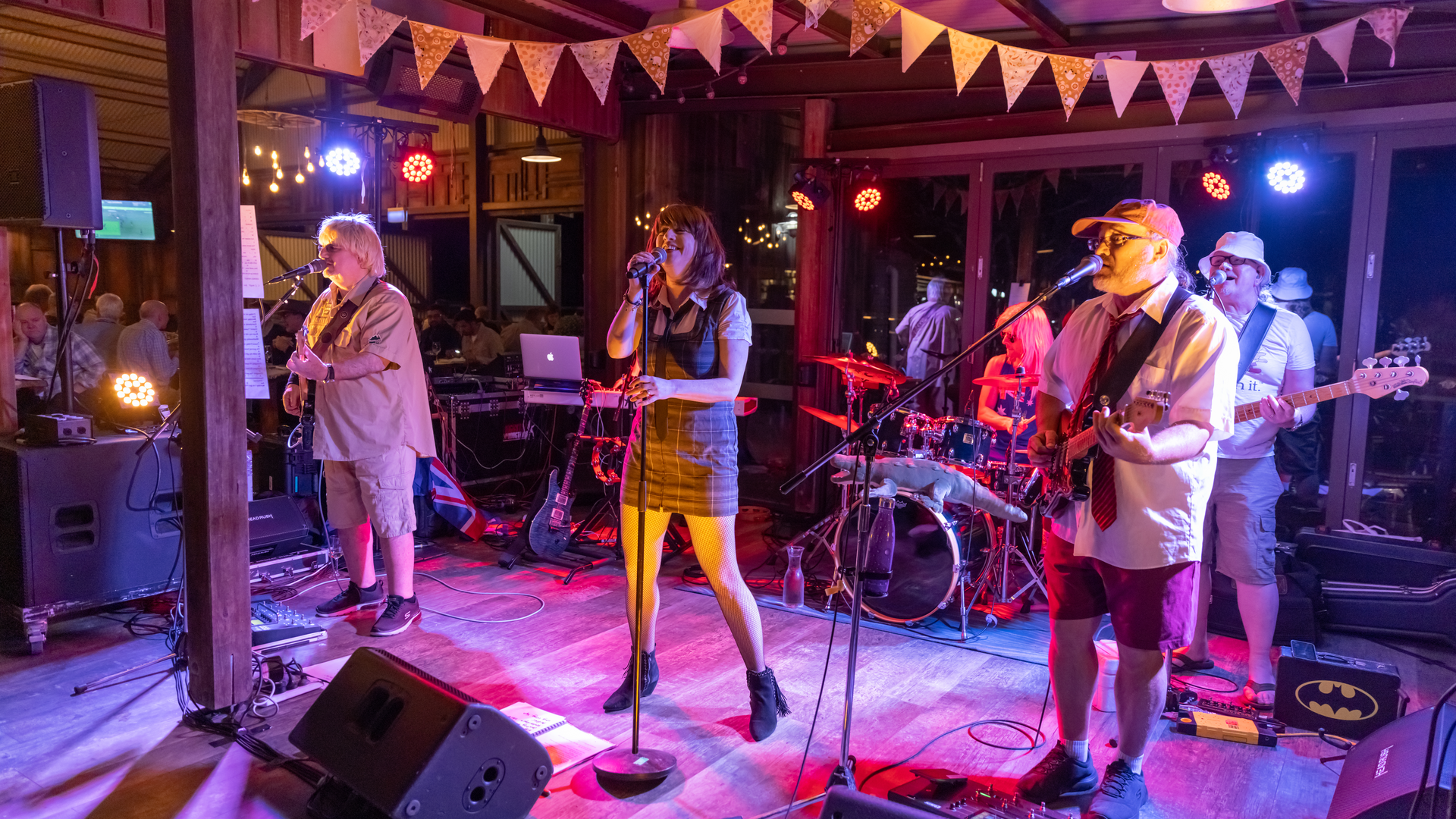 A five-member band performs on a decorated indoor stage with instruments including guitar, bass, drums, and keyboard, while colorful lights illuminate the scene.