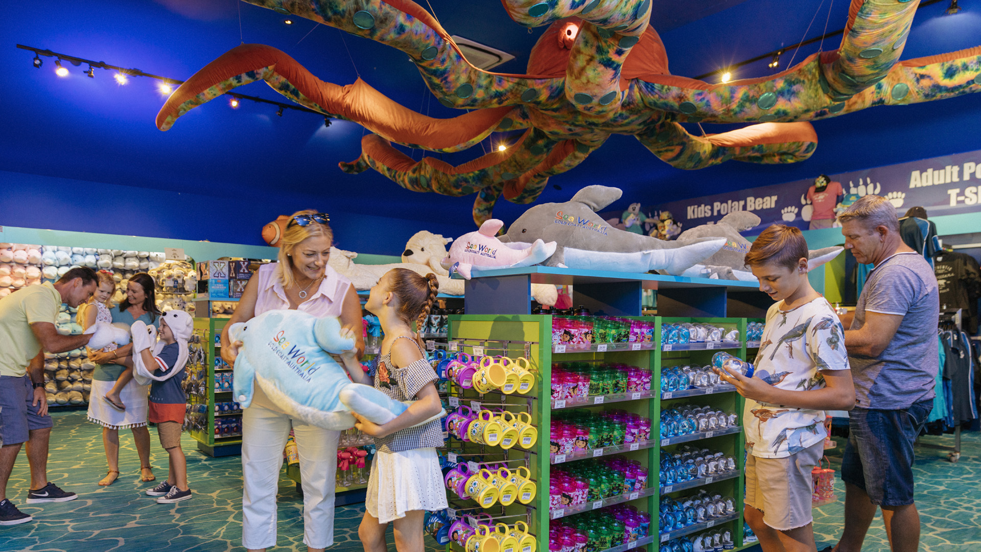 People shop for stuffed animals and souvenirs in a brightly lit, ocean-themed gift shop with a large octopus decoration hanging from the ceiling.