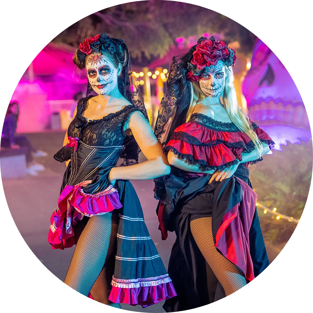Two women in colorful Day of the Dead costumes and face paint pose together outdoors at night, surrounded by festive lights and decorations.